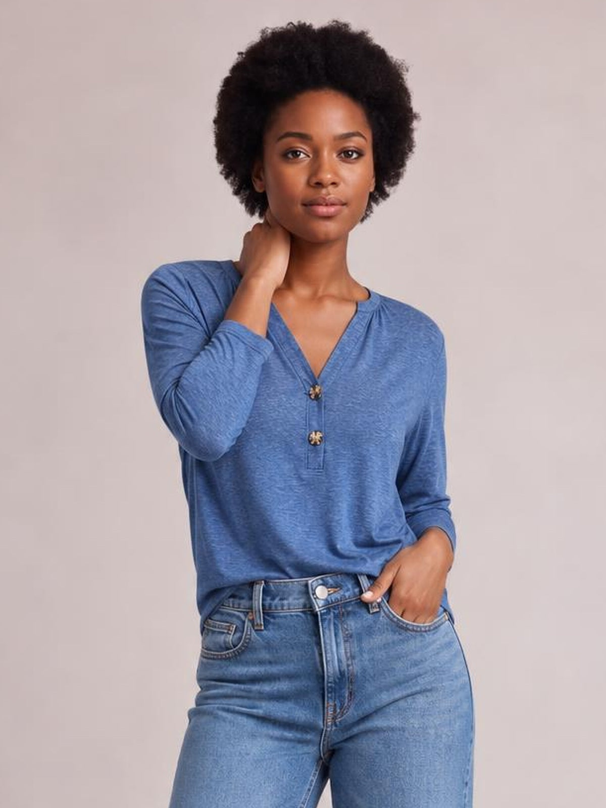 A model wears a blue notched long sleeve T-shirt with a relaxed silhouette, V-shaped neckline, and button front, touching her neck and resting a hand in her pocket.