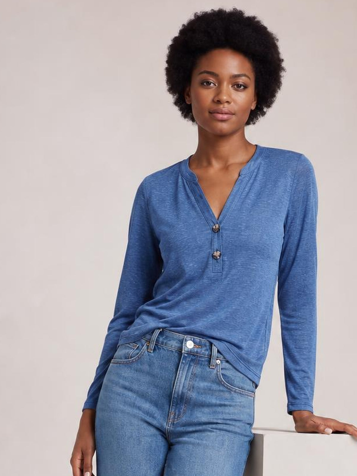 Model wearing a blue notched long sleeve T-shirt with a relaxed silhouette, V-neckline, and two-button placket, leaning casually with one hand resting nearby.