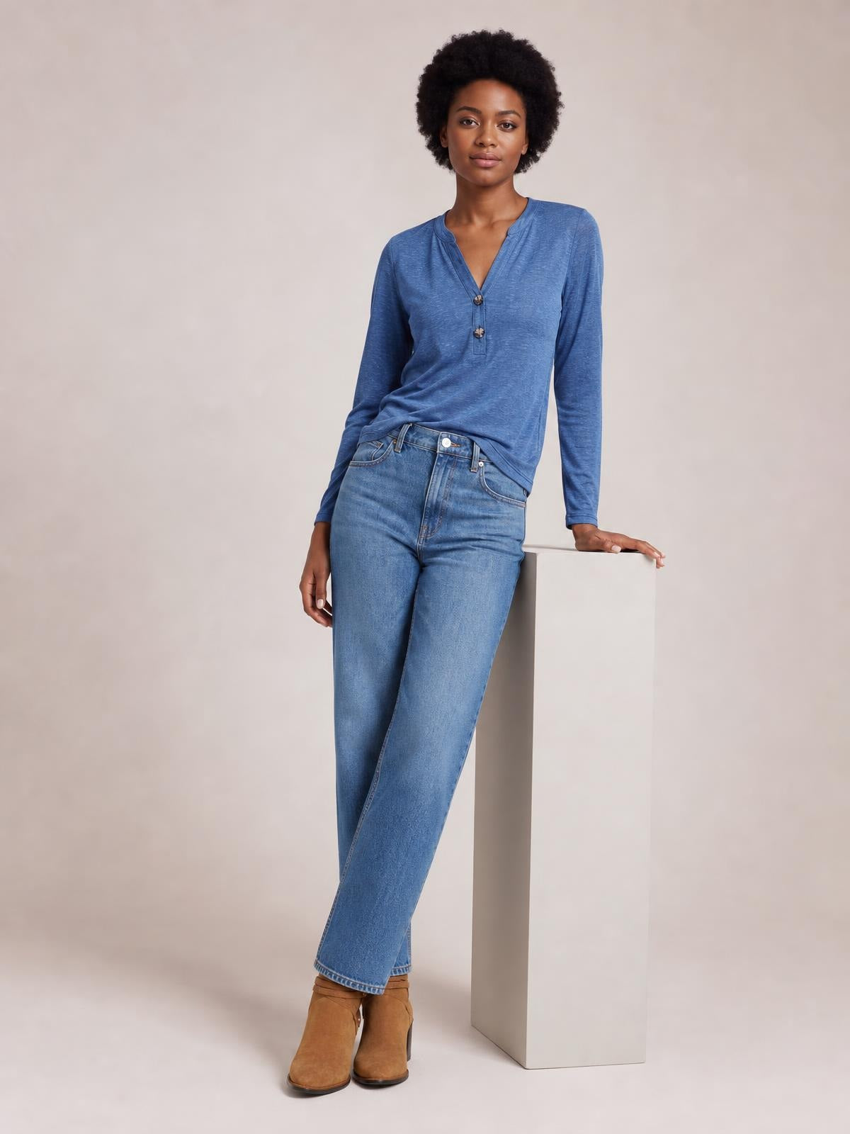 A model wears a blue notched long sleeve T-shirt with a V-neck button placket and relaxed silhouette, leaning casually with one hand on a pedestal.