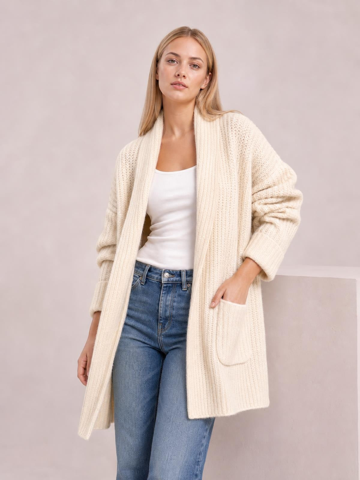 Model wears a cream open-front ribbed cardigan with long sleeves, a relaxed longline silhouette, and large front pockets over a white top and jeans.