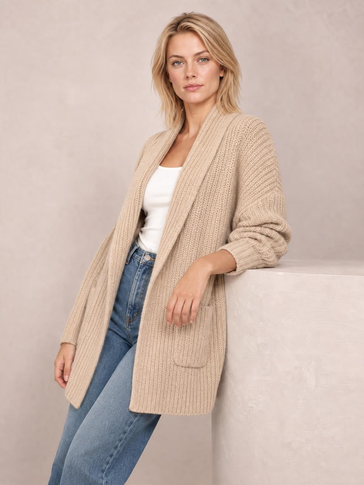 A model leans against a ledge wearing a beige ribbed open-front cardigan with long sleeves, a relaxed longline silhouette, and front patch pockets.