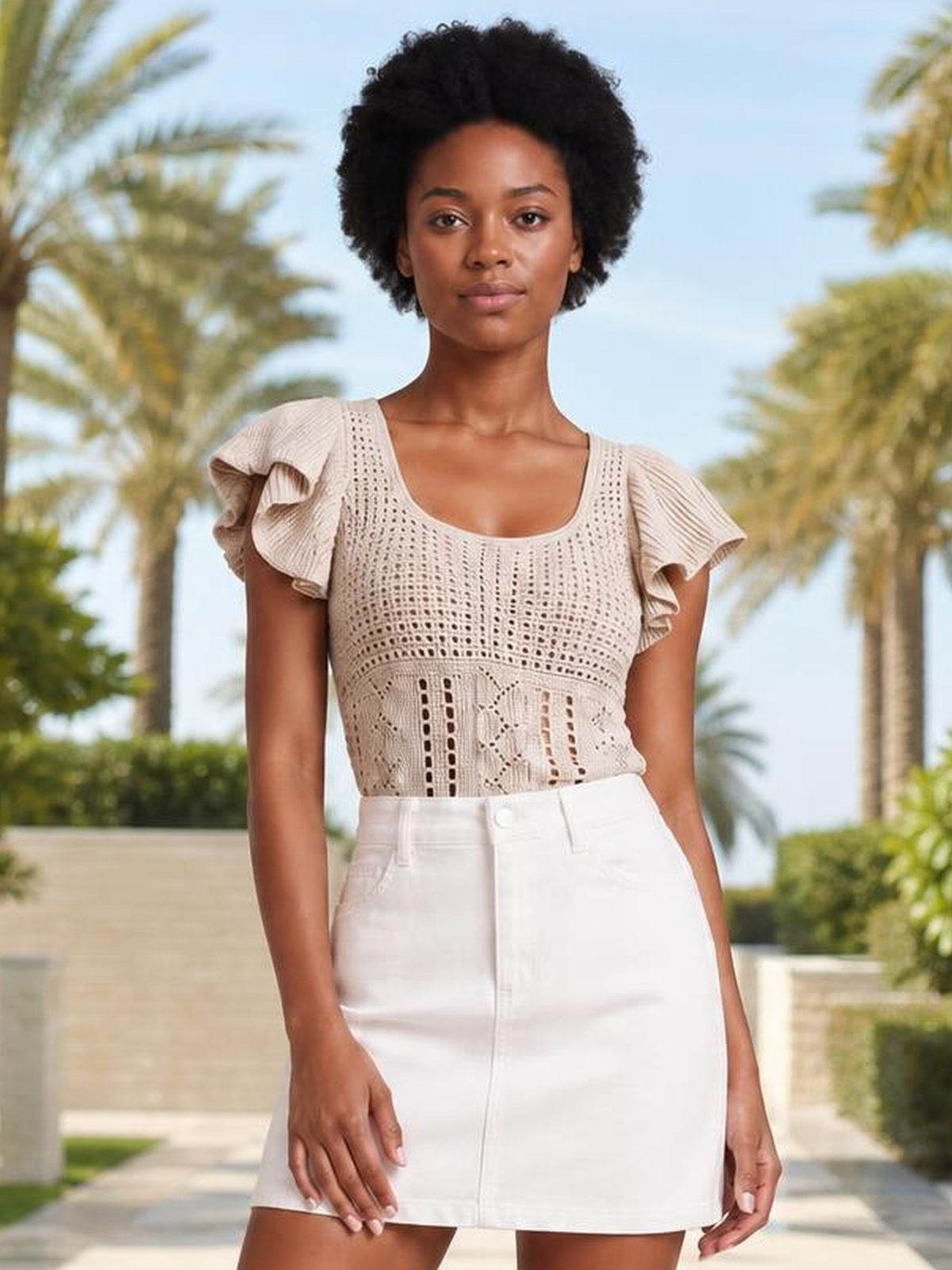 A model stands wearing a beige openwork knit top with a square neckline, flutter sleeves, and a fitted silhouette, paired with a white high-waisted skirt.