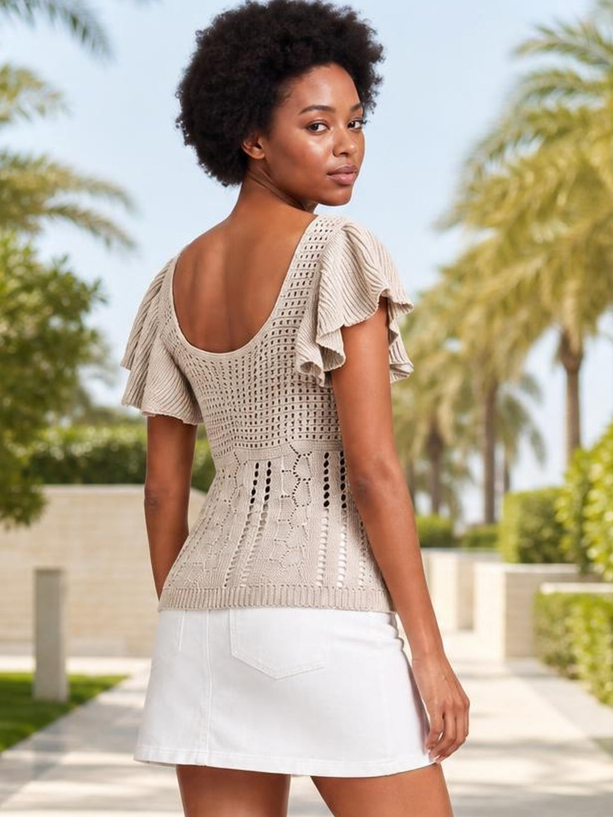 Model turns to show a beige openwork knit top with layered flutter sleeves, a scoop back, fitted silhouette, and ribbed hem.