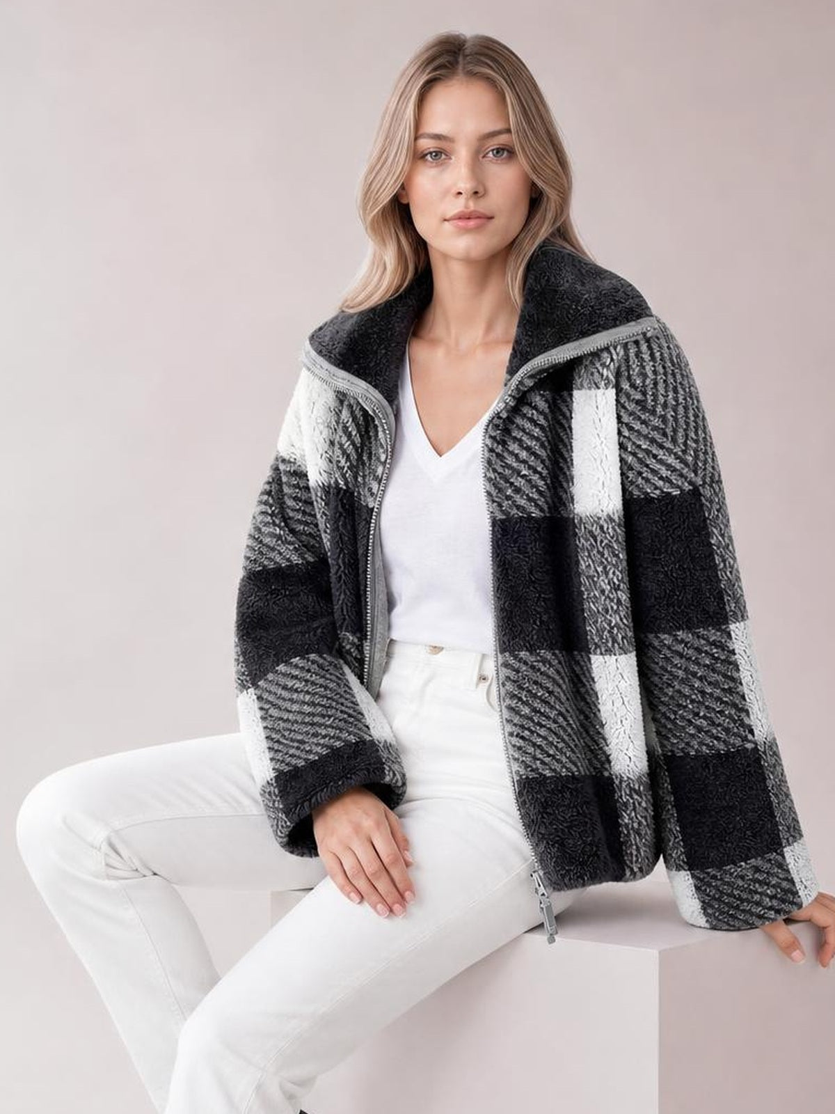 A seated model wears a black, gray, and white plaid zip-up jacket with long sleeves, a high collar, textured panels, and a relaxed oversized silhouette.