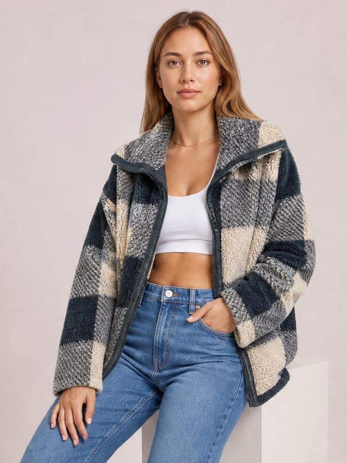 A seated model wears an oversized gray, black, and cream plaid zip-up jacket with long sleeves, a wide collar, and a relaxed silhouette.