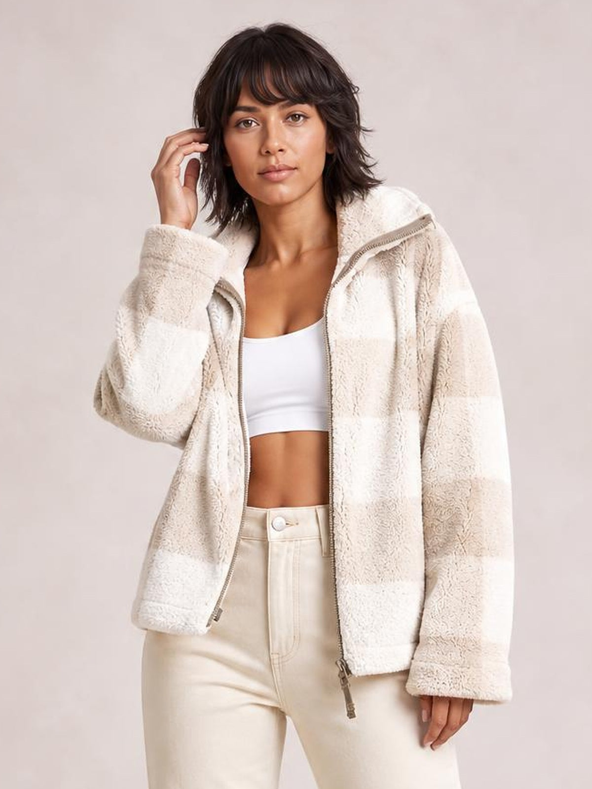 Model wears a cream zip-up jacket with long sleeves, a high collar, oversized silhouette, textured paneling, and a front zipper, posing with one hand near her hair.