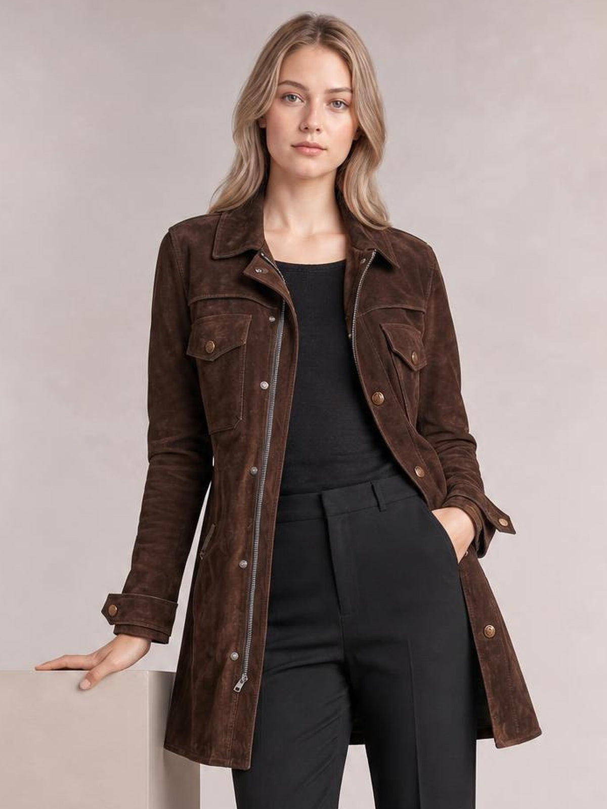 Model poses in a brown long-sleeve jacket with a collared front, zip closure, flap pockets, button details, and a long straight silhouette.