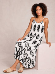 Model seated in a flowy white cami dress with slim spaghetti straps, a V-neckline, bold black geometric print, and an ankle-grazing silhouette.