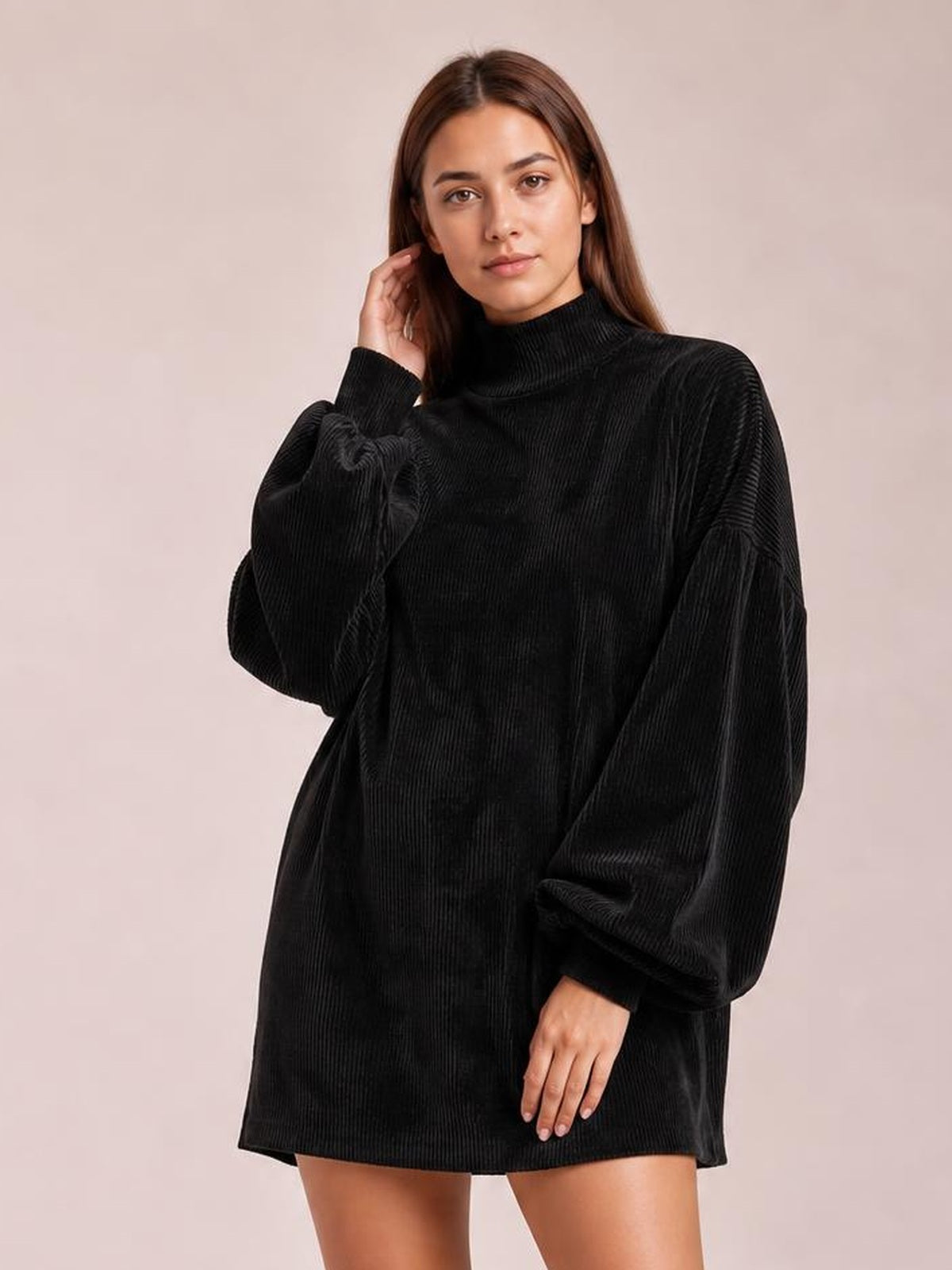 Model poses with one hand near her hair in a black ribbed mini dress featuring a mock neck, relaxed silhouette, and voluminous lantern sleeves.