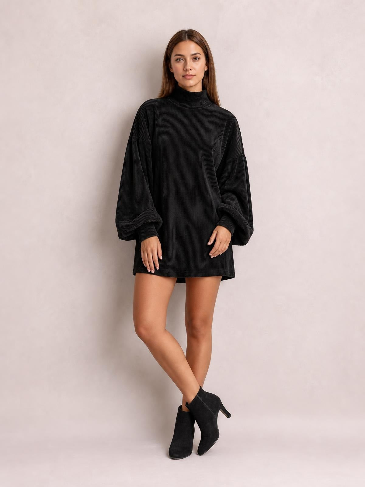 Model wears a black ribbed mini dress with a mock neck, oversized lantern sleeves, relaxed straight silhouette, and rolled cuffs while standing with crossed legs.