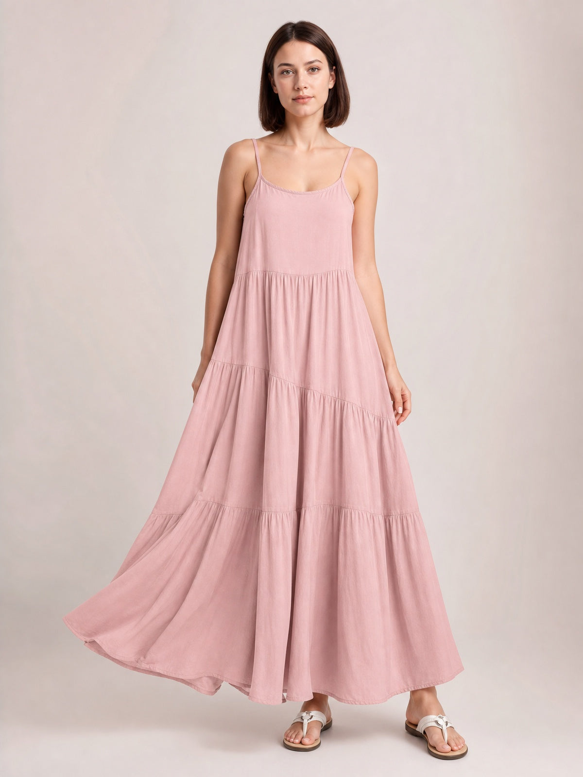 Model poses in a flowing blush pink ruched tiered spaghetti strap maxi dress with a scoop neckline, gathered seams, and full floor-skimming skirt.