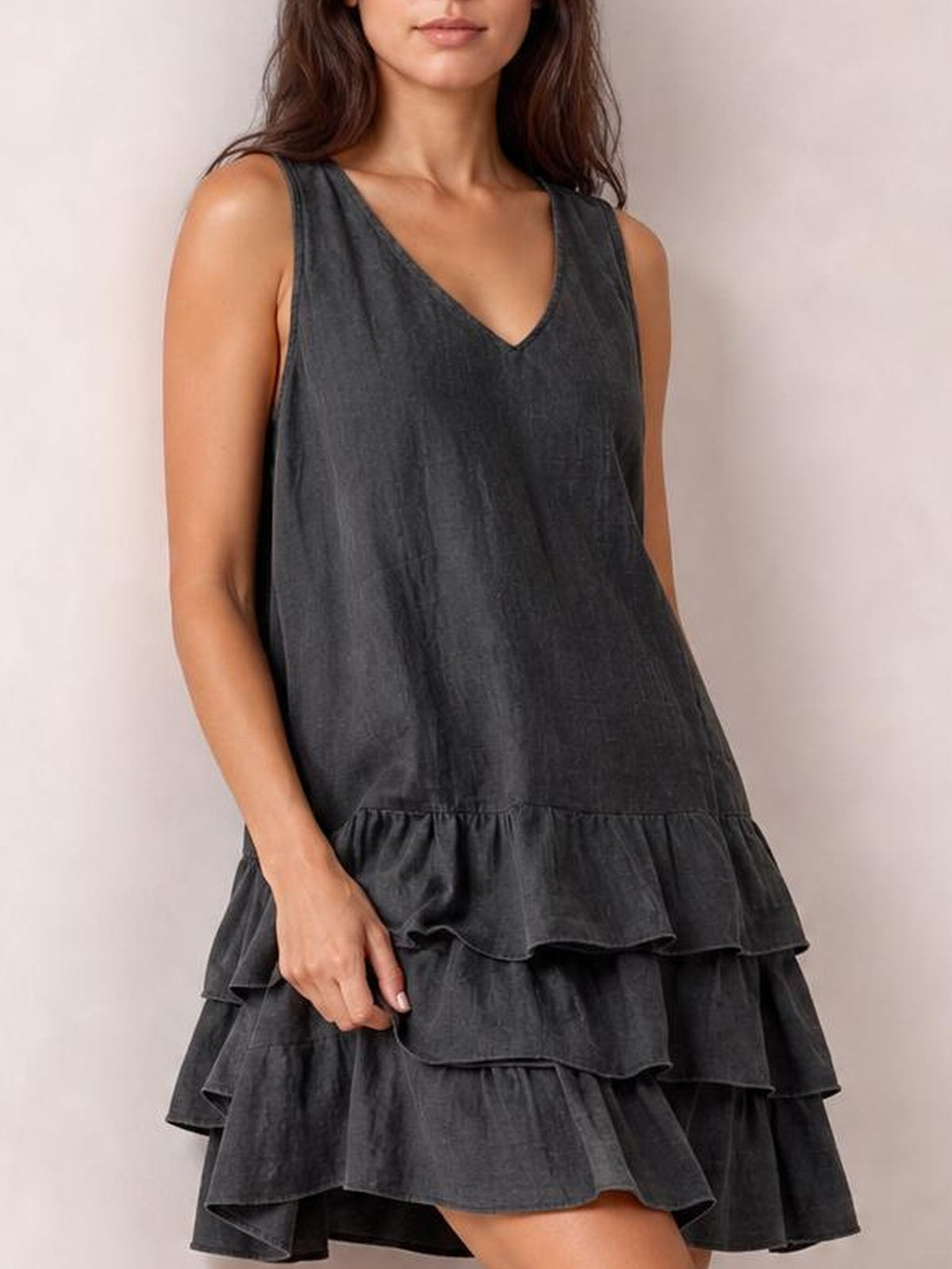 A model wears a charcoal gray plus-size sleeveless V-neck dress with a loose tiered silhouette and multiple ruffled skirt layers, lightly holding the hem.