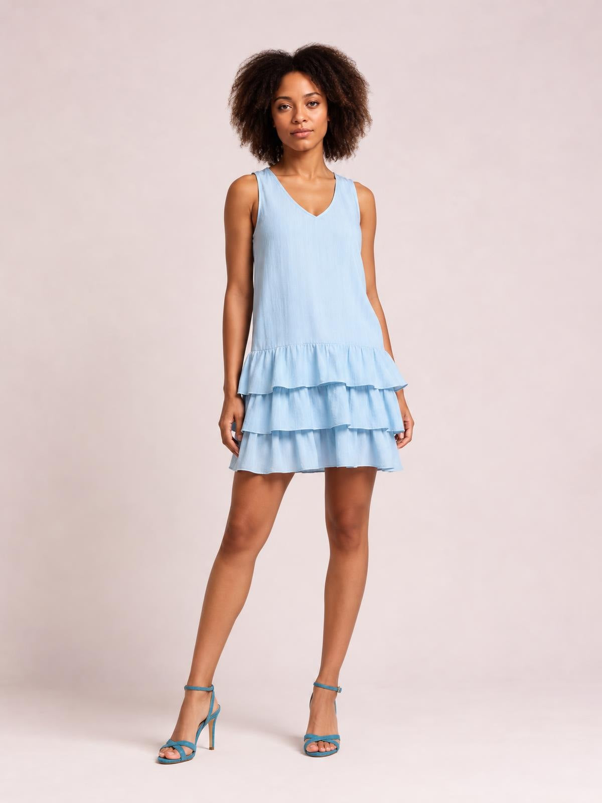 A model stands with one leg crossed in a light blue plus-size sleeveless dress featuring a V-neck, loose silhouette, and tiered ruffled hem.
