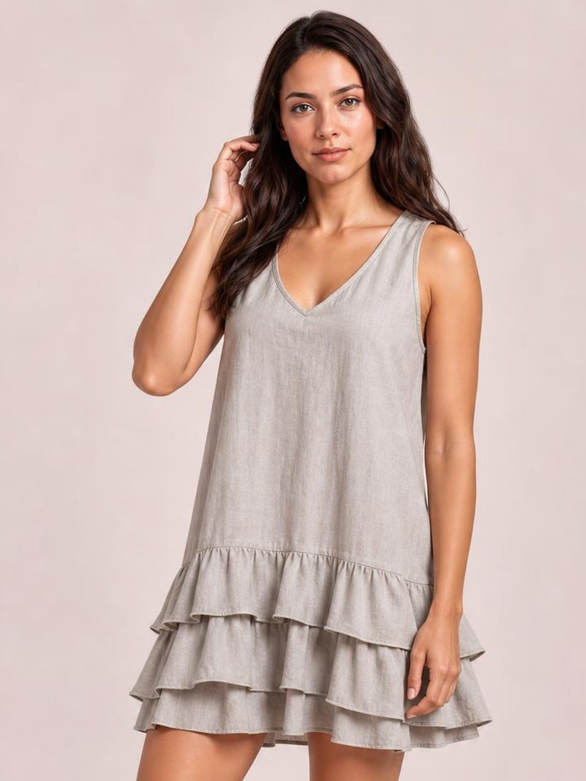 A model wears a light gray sleeveless V-neck dress with a relaxed silhouette, tiered construction, and multiple ruffled hem layers, touching her hair.