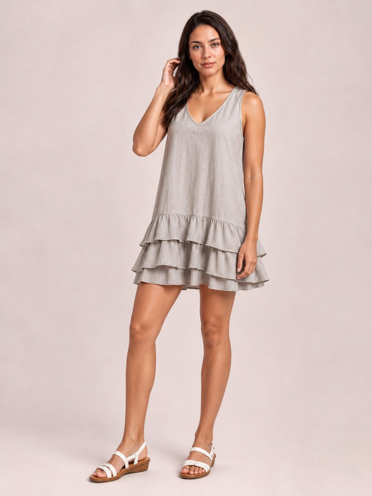 A model poses in a light gray sleeveless V-neck dress with a relaxed shift silhouette and tiered ruffled hem, touching her hair with one hand.