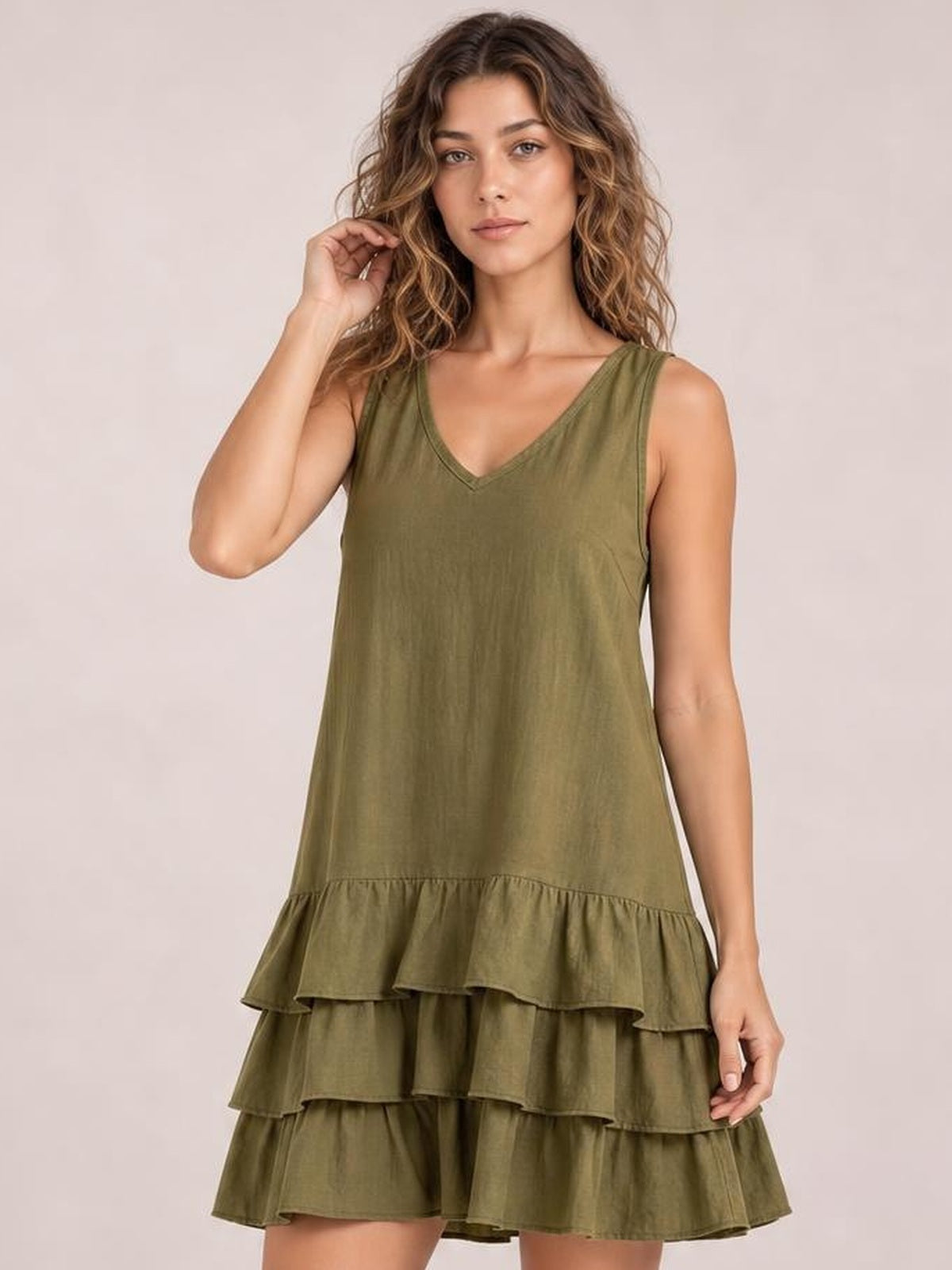 Model poses in an olive green sleeveless V-neck dress with a relaxed A-line silhouette and tiered ruffled hem, touching her hair.