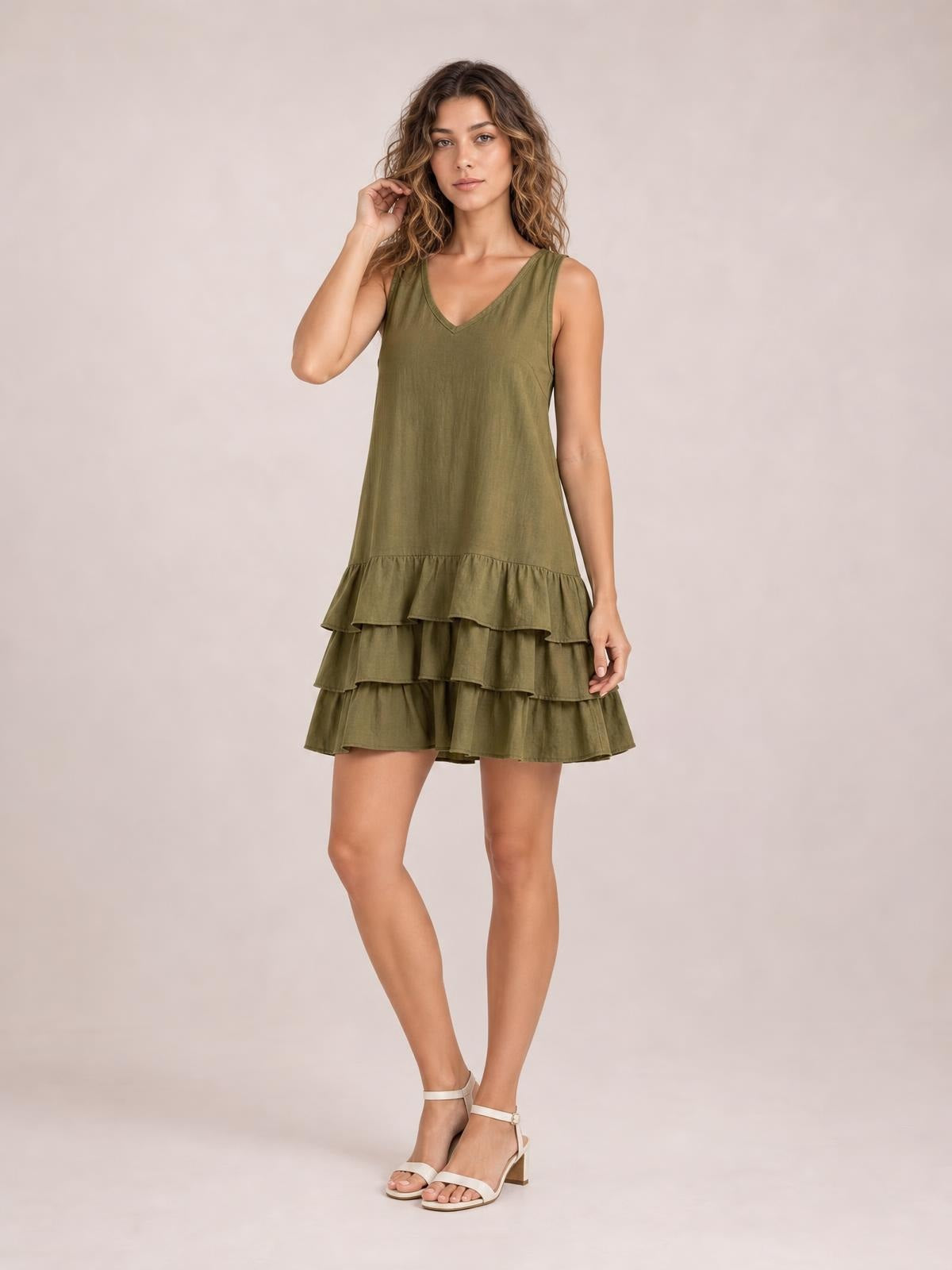 A model poses in an olive green sleeveless V-neck dress with a relaxed silhouette and tiered ruffled hem, touching her hair with one leg crossed.