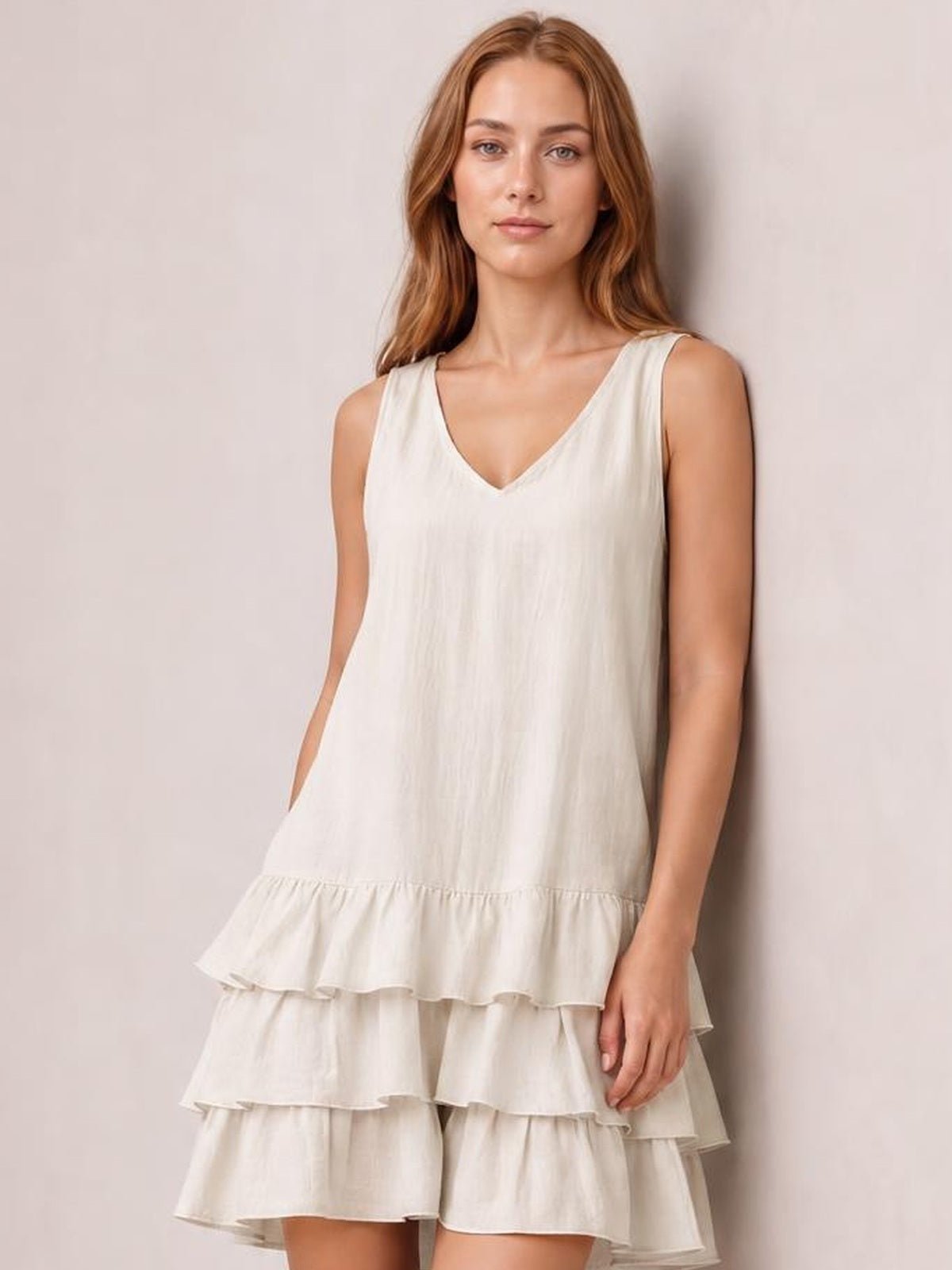Model poses in a cream sleeveless V-neck dress with a relaxed silhouette, gathered waist seam, and tiered ruffled hem.