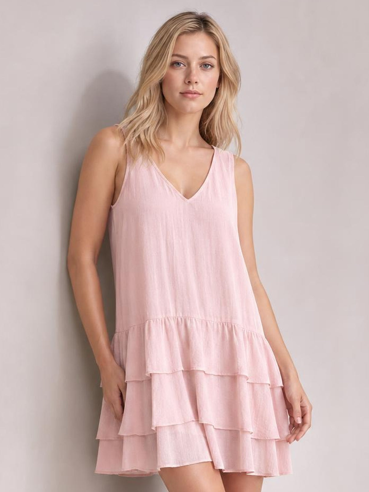 A model poses in a pale pink sleeveless V-neck dress with a relaxed A-line silhouette and multiple tiered ruffled hems, holding the skirt at one side.