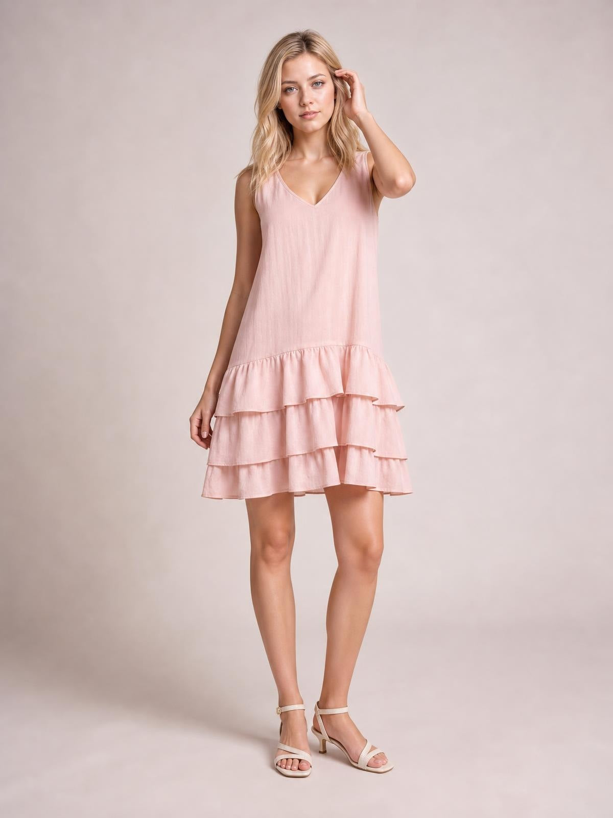 A model stands in a blush pink sleeveless dress with a V-neck, relaxed silhouette, and tiered ruffled hem, touching her hair with one hand.