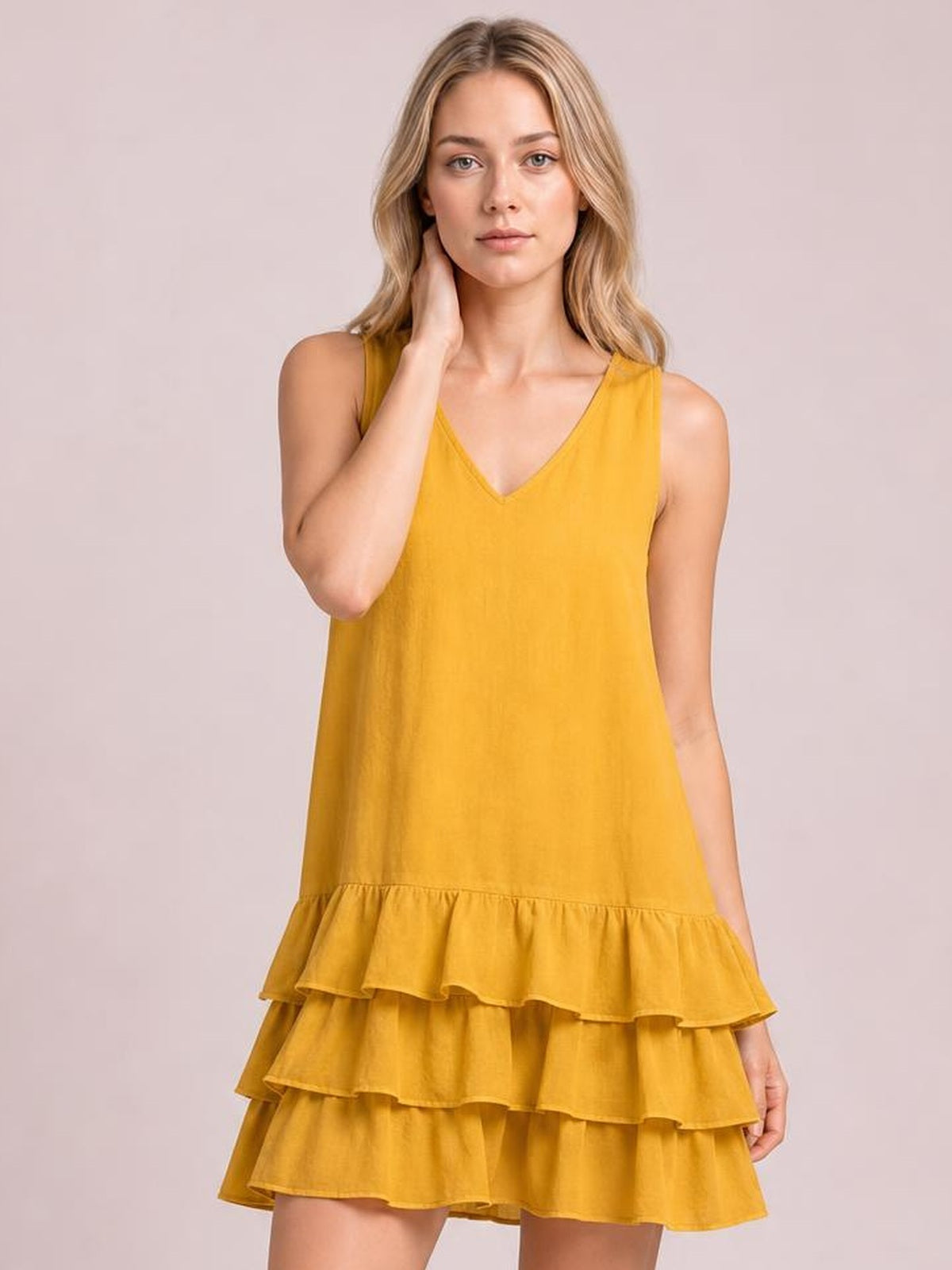 A model poses in a mustard-yellow sleeveless V-neck dress with a relaxed A-line silhouette and multiple tiered ruffled hem layers, touching her neck.