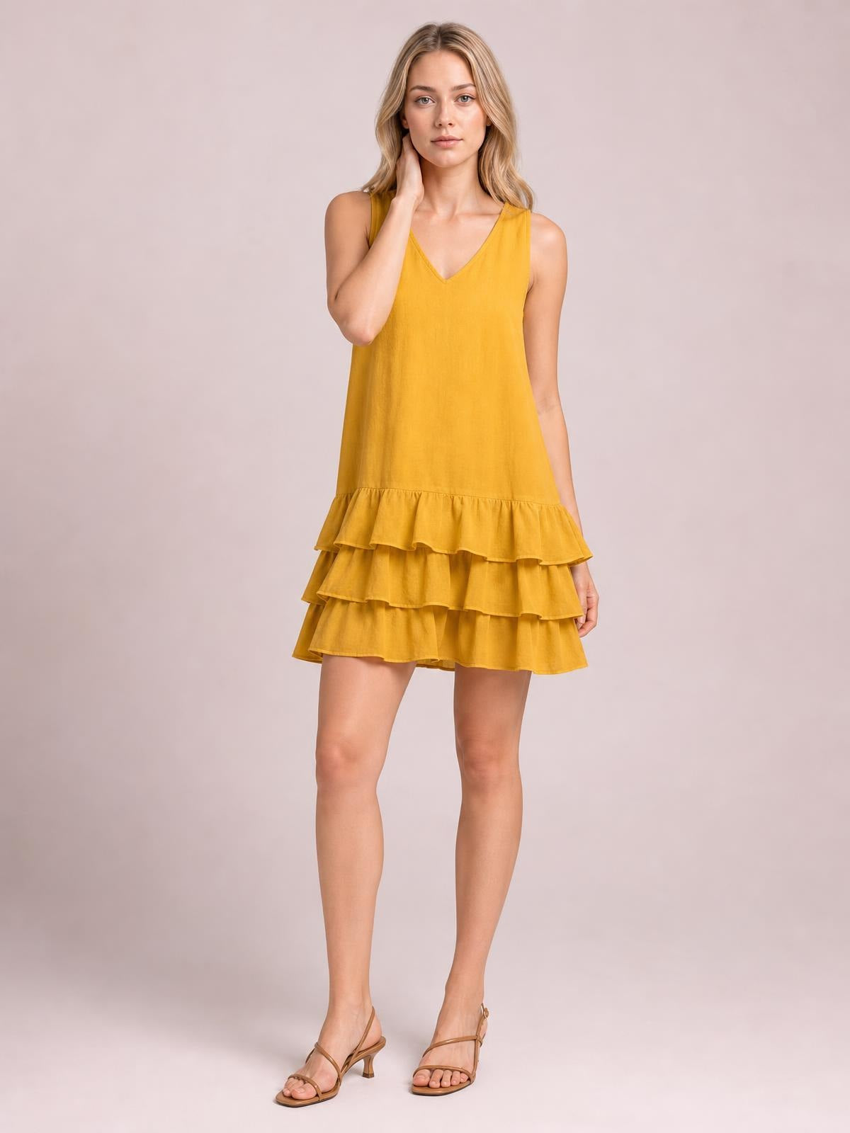 A model poses in a mustard-yellow sleeveless V-neck dress with a relaxed silhouette and tiered ruffled hem, touching her neck with one hand.