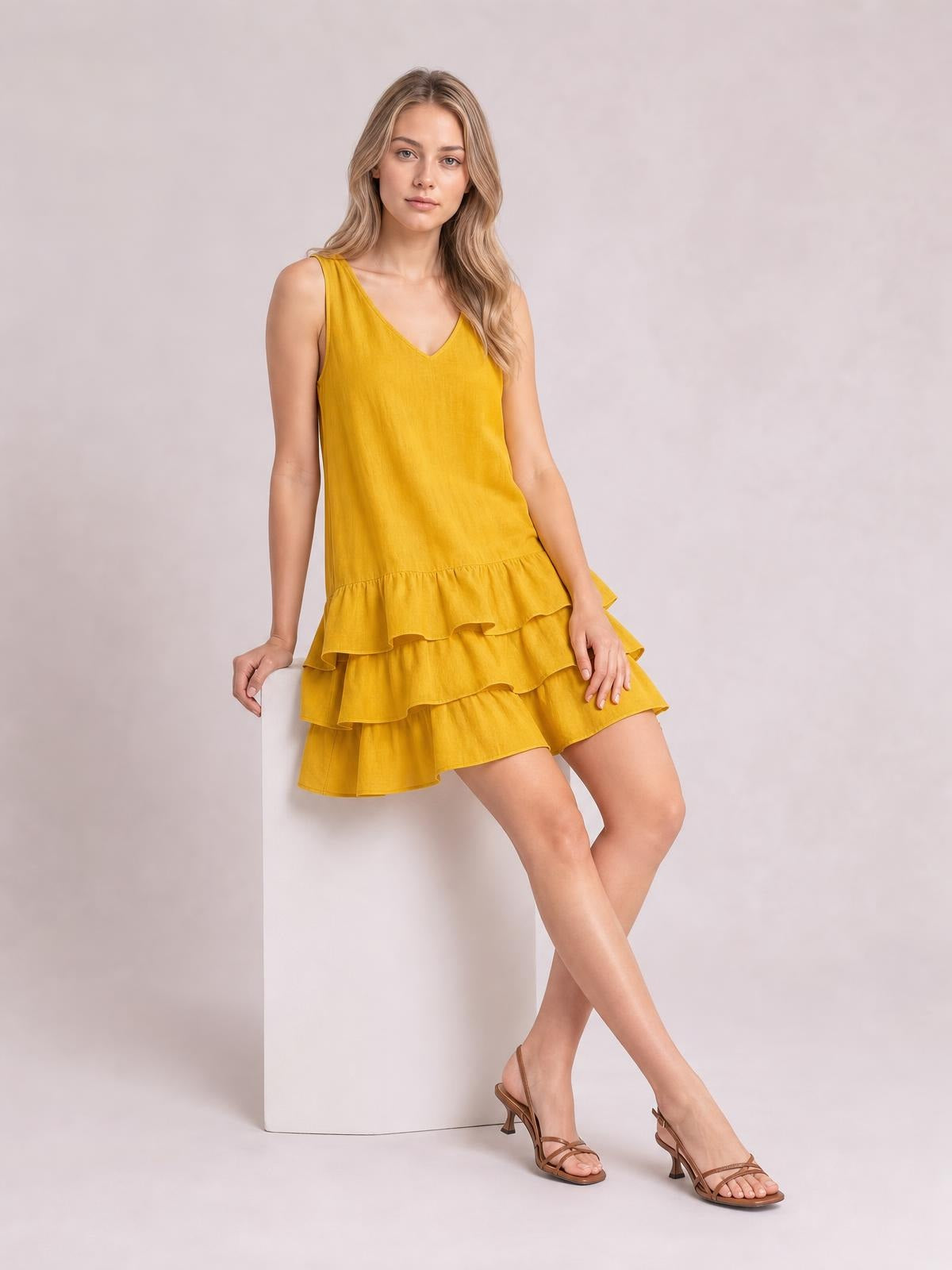 A model sits with crossed legs in a mustard yellow sleeveless V-neck dress featuring a relaxed silhouette and three tiered ruffled hems.