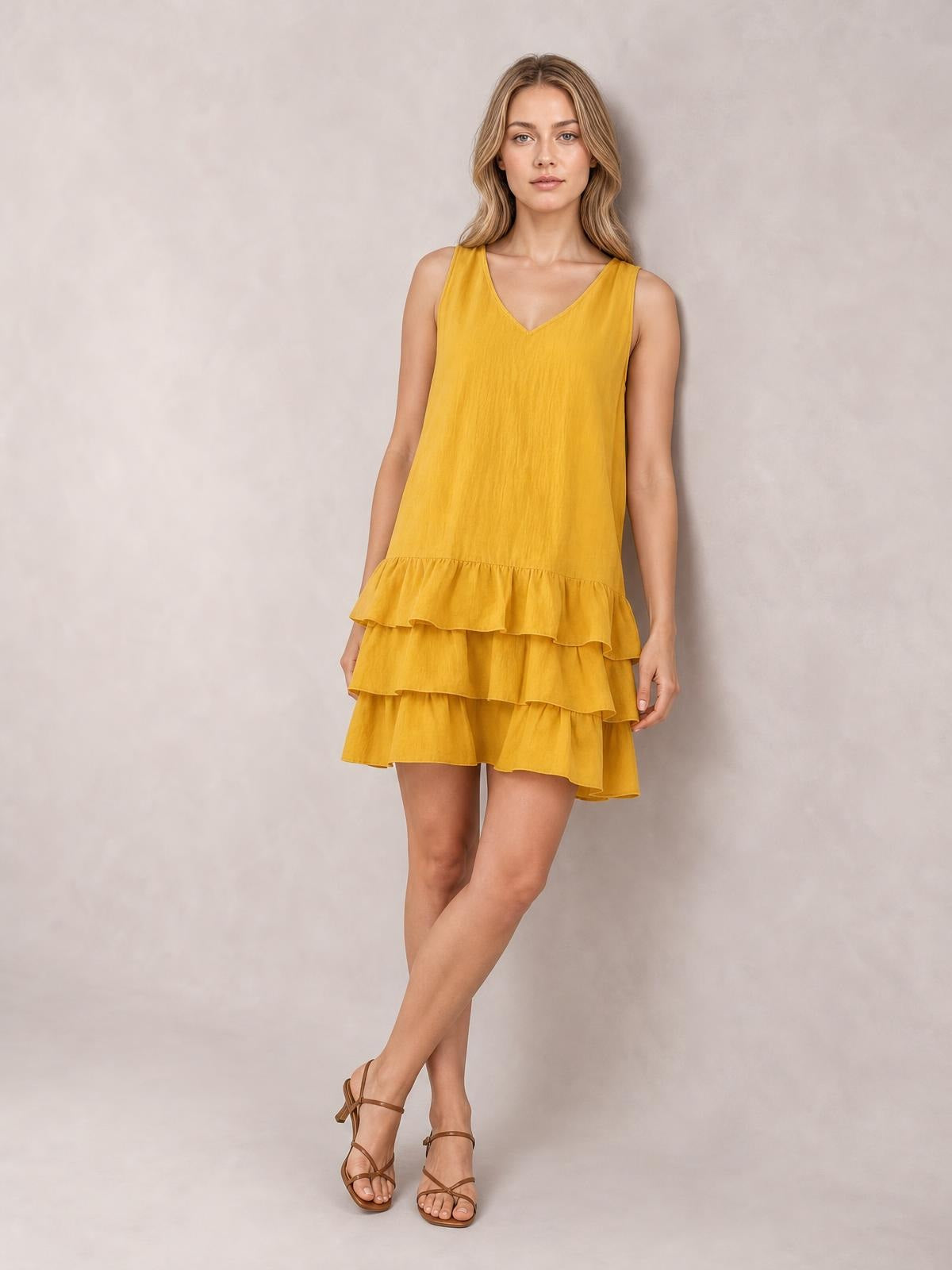 Model wears a mustard-yellow sleeveless V-neck dress with a relaxed silhouette and three tiered ruffled hems, standing with crossed legs.