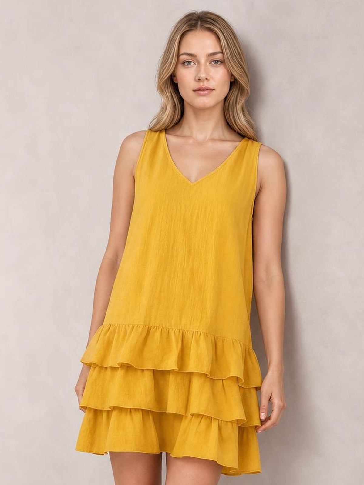 Model poses in a mustard yellow sleeveless V-neck dress with a relaxed silhouette, tiered construction, and multiple ruffled hems.