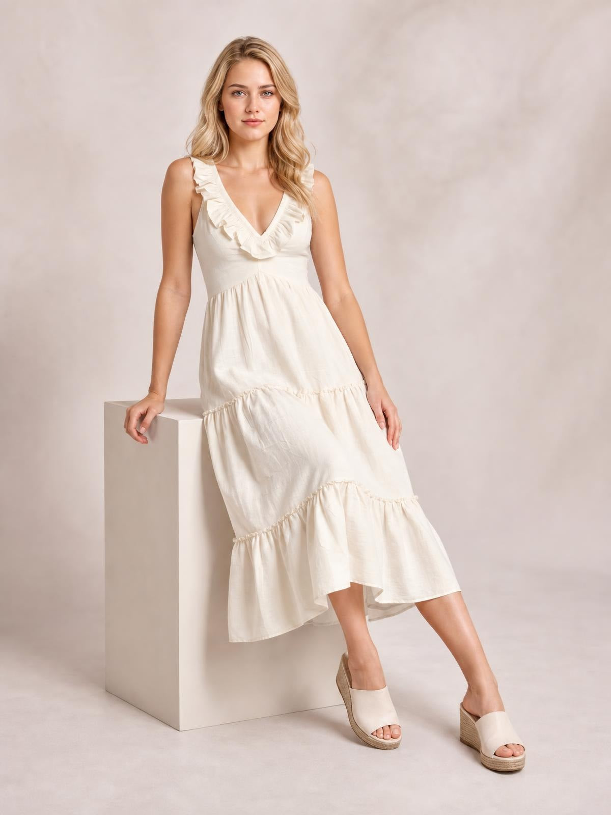 Model poses in a cream ruffled V-neck tiered midi dress with a gently fitted bodice, flowing tiered skirt, and sleeveless silhouette.