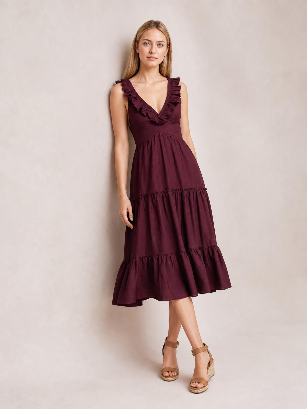 Model poses in a burgundy sleeveless midi dress with a deep V-neck, ruffled shoulder trim, gathered waist, and flowing tiered silhouette.