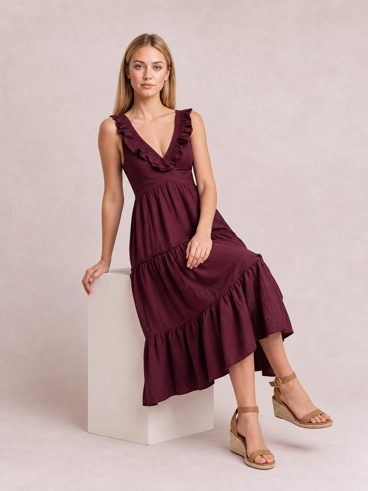 Model seated in a burgundy ruffled V-neck midi dress with a gathered waist, tiered full skirt, and flowing silhouette.