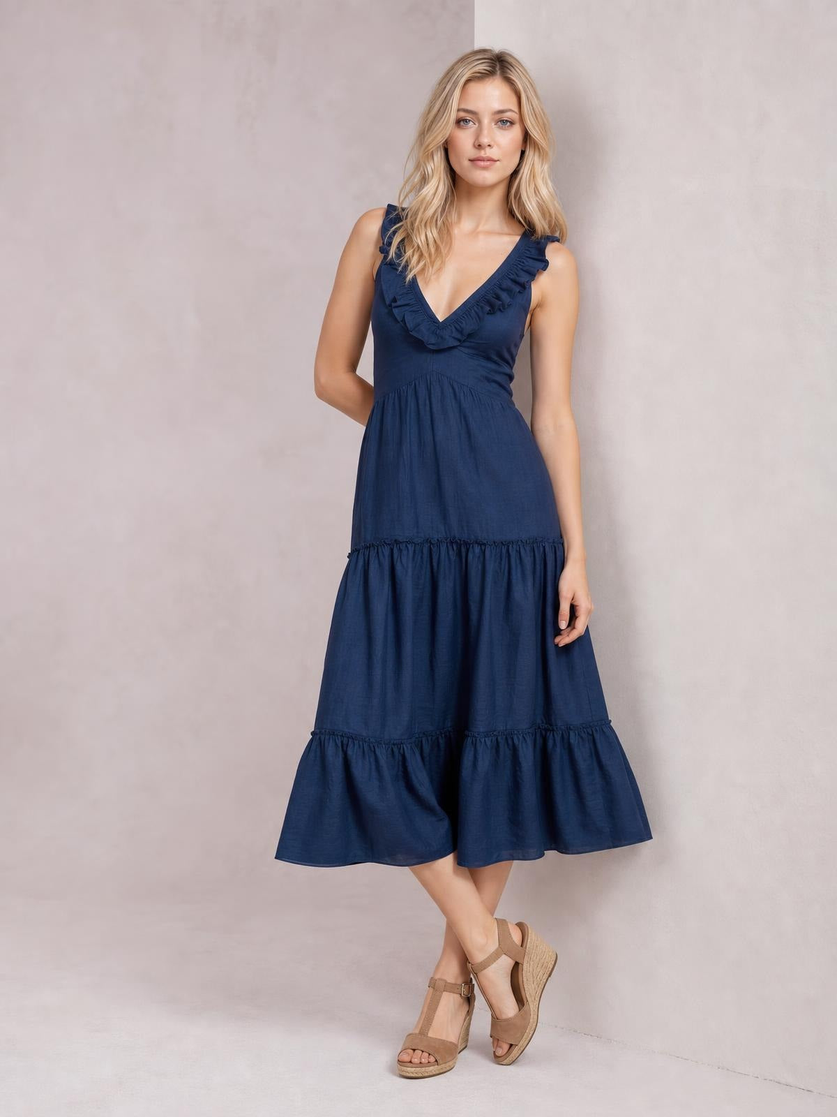 A model poses in a navy sleeveless midi dress with a ruffled V-neck, fitted bodice, gathered waist, and flowing tiered skirt.
