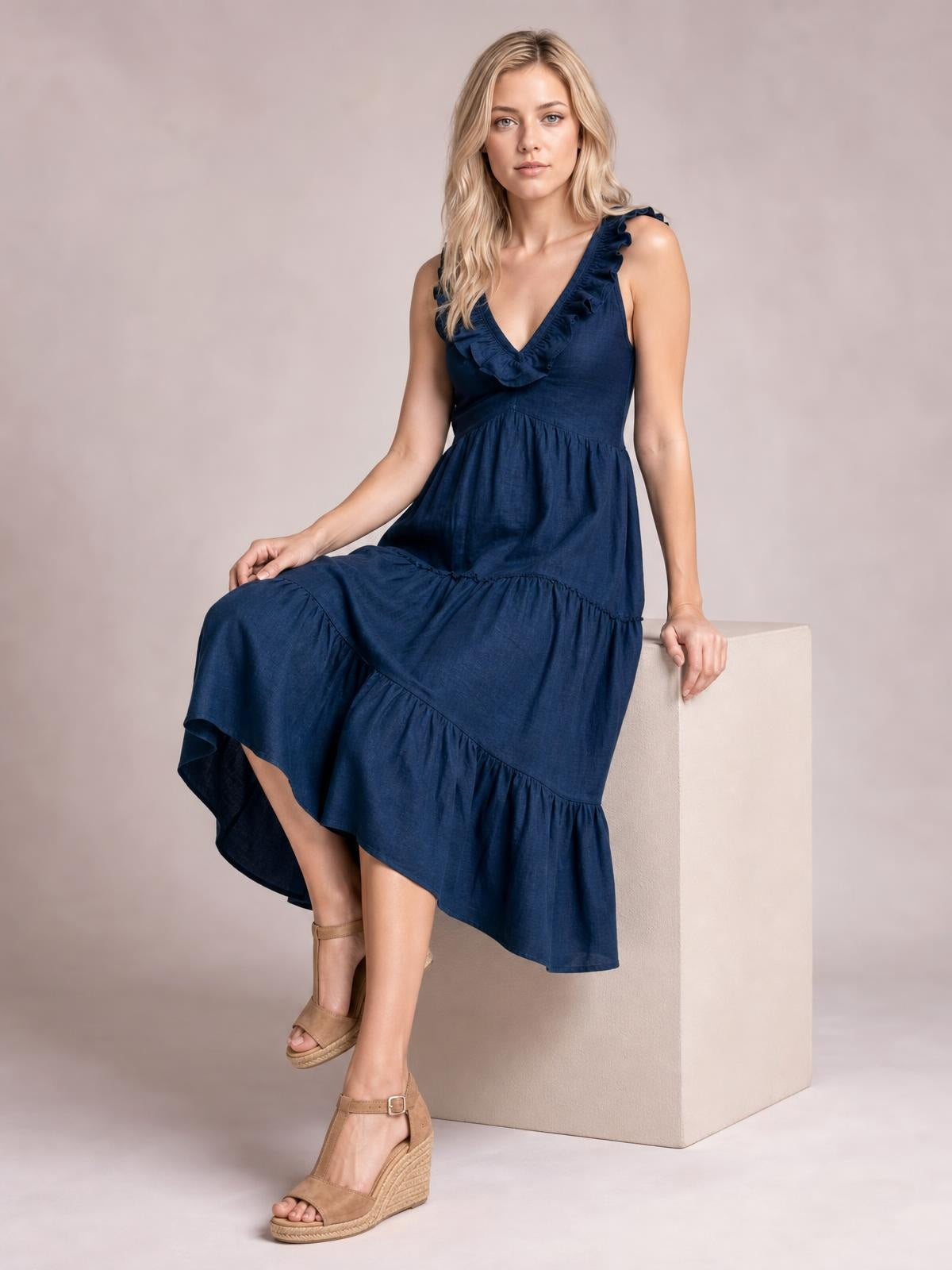 A model sits in a navy ruffled V-neck midi dress with sleeveless styling, a gathered waist, tiered skirt, and flowing A-line silhouette.