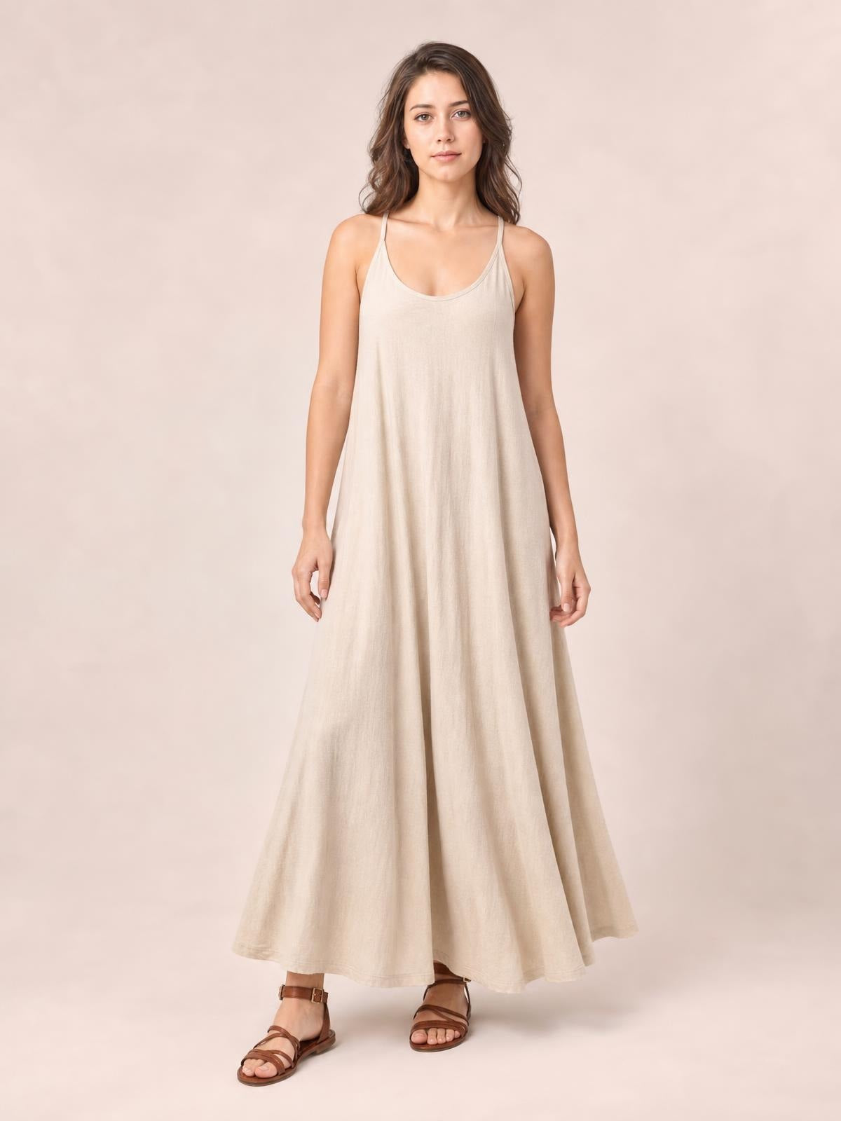 A model stands in a flowing beige scoop-neck maxi dress with thin spaghetti straps, a relaxed A-line silhouette, and softly draped construction.