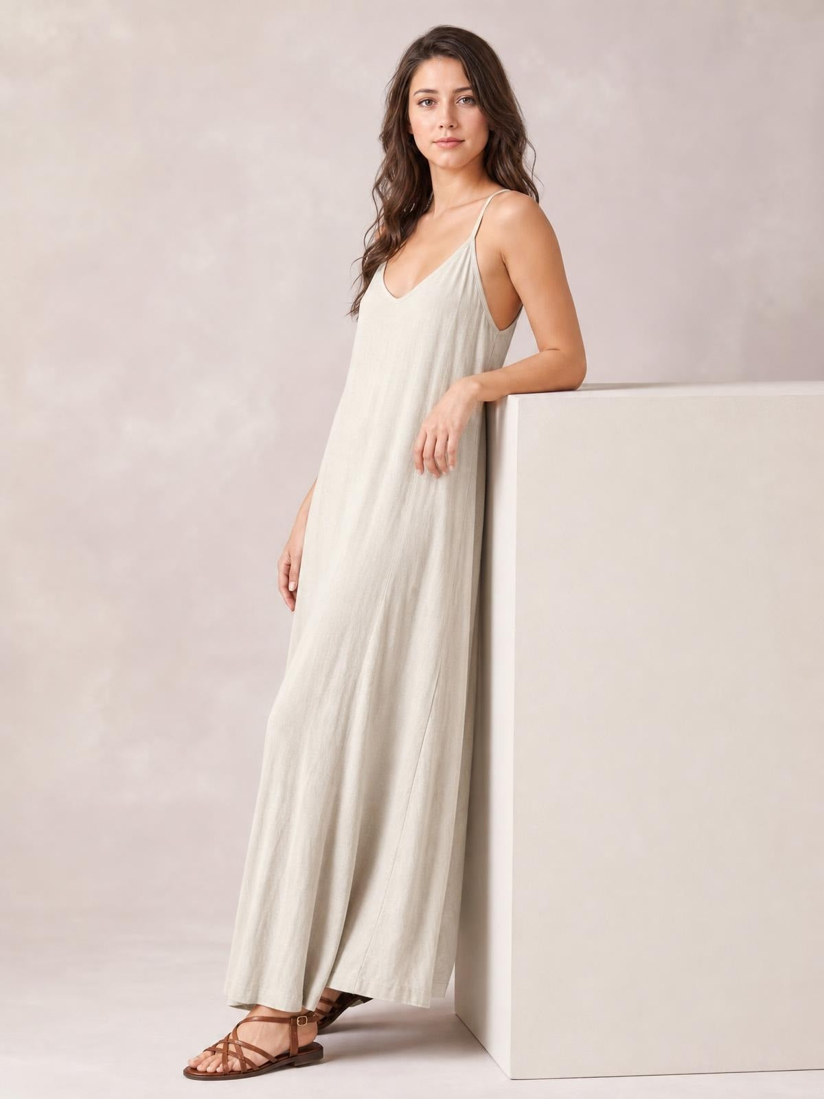 A model leans against a ledge wearing a light beige scoop-neck maxi dress with spaghetti straps and a relaxed, flowing silhouette.