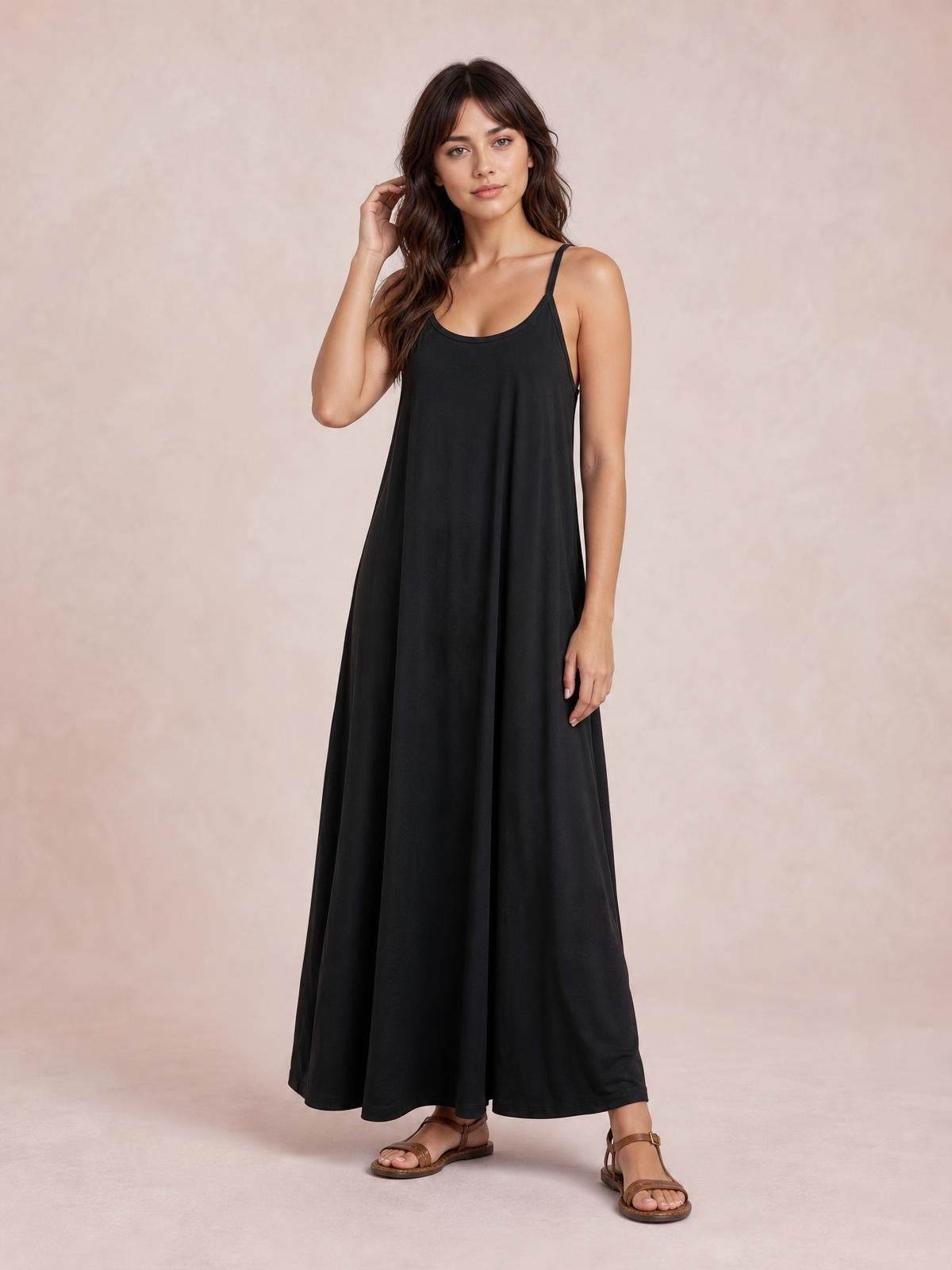 Model poses with one hand in her hair wearing a black scoop-neck spaghetti-strap maxi dress with a loose, flowing floor-length silhouette.