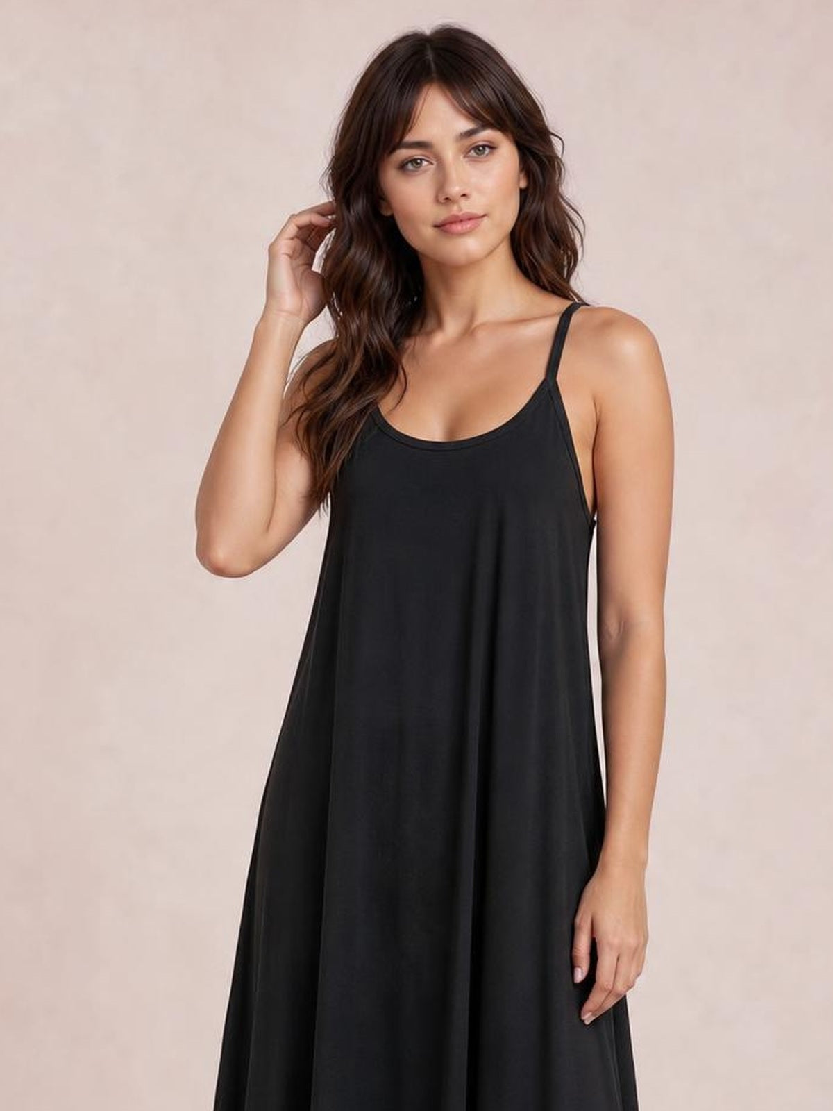 A model wears a black plus-size maxi dress with a scoop neck, thin spaghetti straps, and a loose, flowing silhouette while touching her hair.