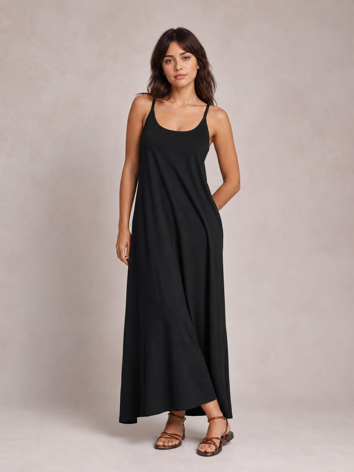 Model poses in a black scoop-neck spaghetti strap maxi dress with a relaxed, flowing silhouette and long hem.
