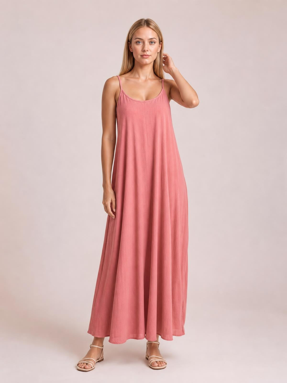 Model wearing a pink scoop neck spaghetti strap maxi dress with a flowing A-line silhouette, standing with one hand touching her hair.