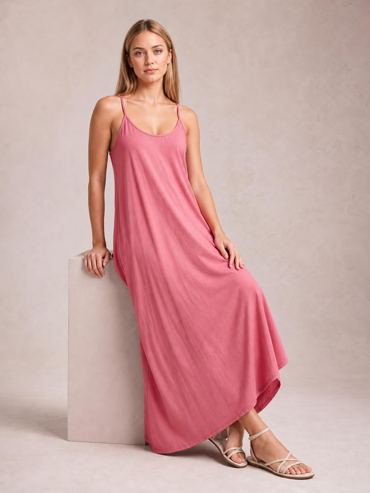 Model seated in a pink scoop-neck maxi dress with thin spaghetti straps, a relaxed flowing silhouette, and visible hem stitching.