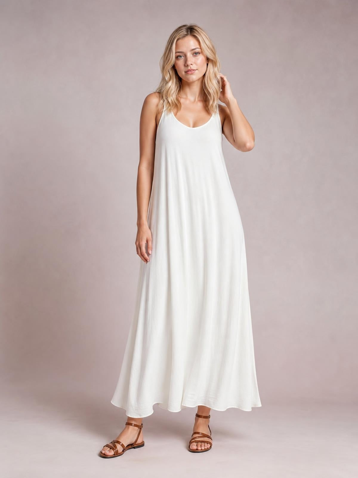 A model poses in a white maxi dress with a scoop neckline, spaghetti straps, and a flowing A-line silhouette, with one hand touching her hair.