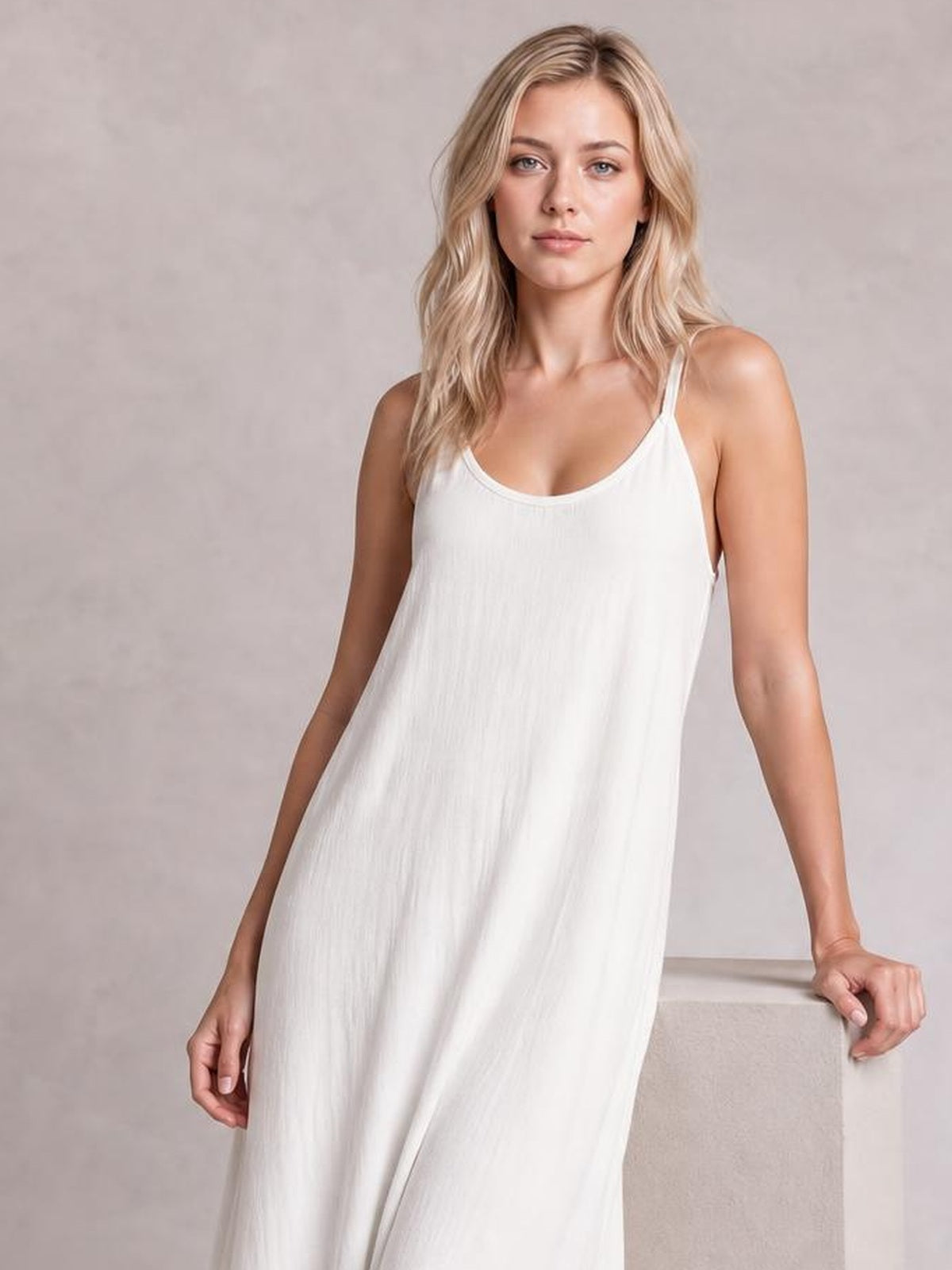 A model wears a white scoop-neck spaghetti-strap maxi dress with a relaxed, flowing silhouette and lightly textured construction while leaning on a ledge.