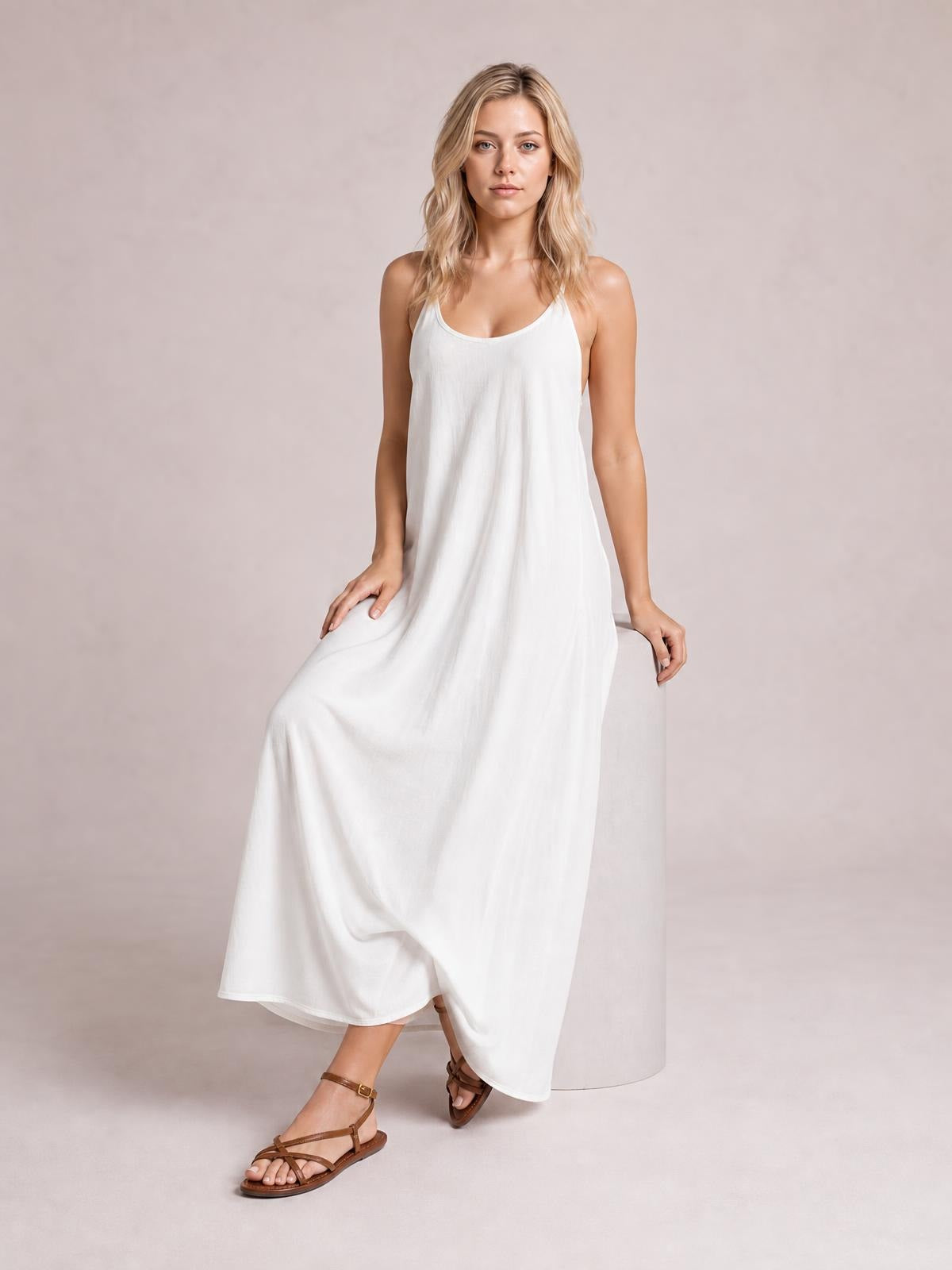 A model sits in a flowing white scoop-neck maxi dress with thin spaghetti straps and a relaxed, full-length silhouette.