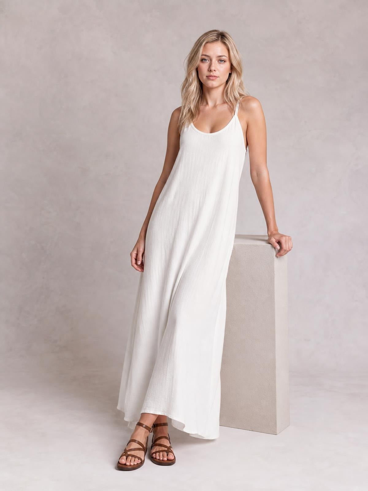 A model wears a white scoop-neck spaghetti-strap maxi dress with a relaxed, flowing silhouette and full-length hem while resting one hand on a block.