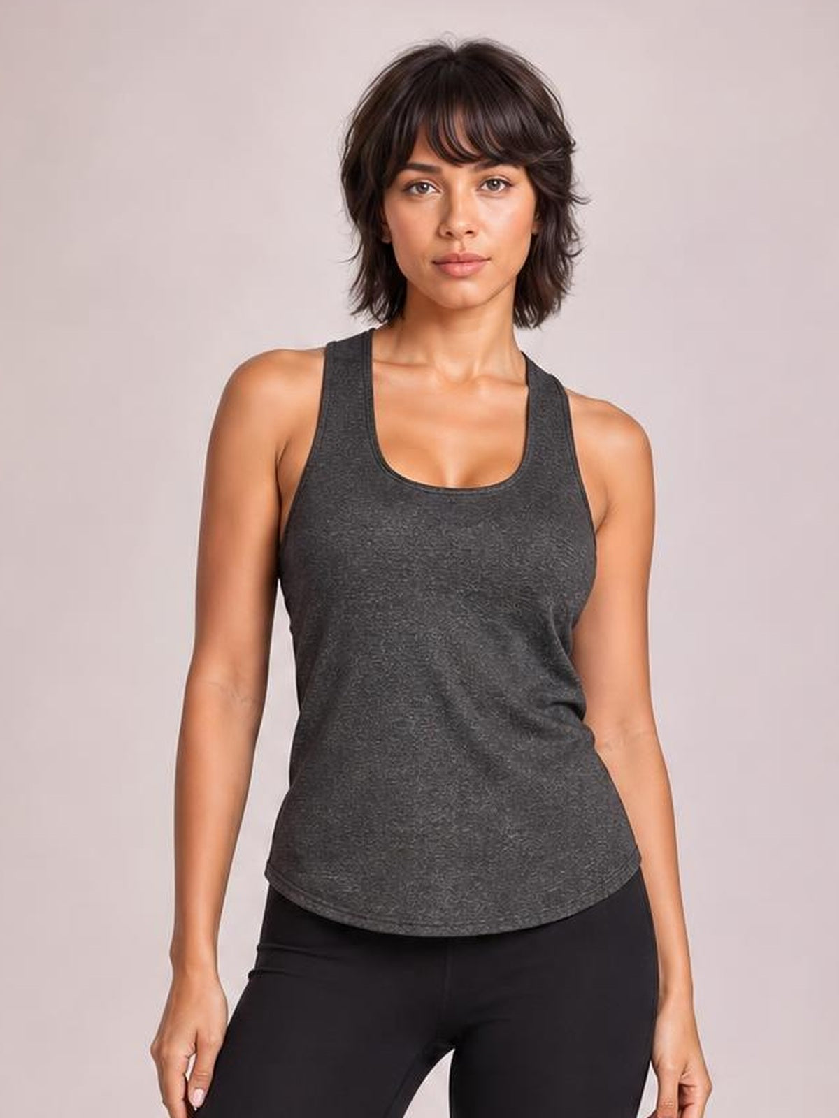 Model wears a charcoal gray scoop neck active tank with wide straps, a relaxed silhouette, and curved hem, standing front-facing with arms at her sides.