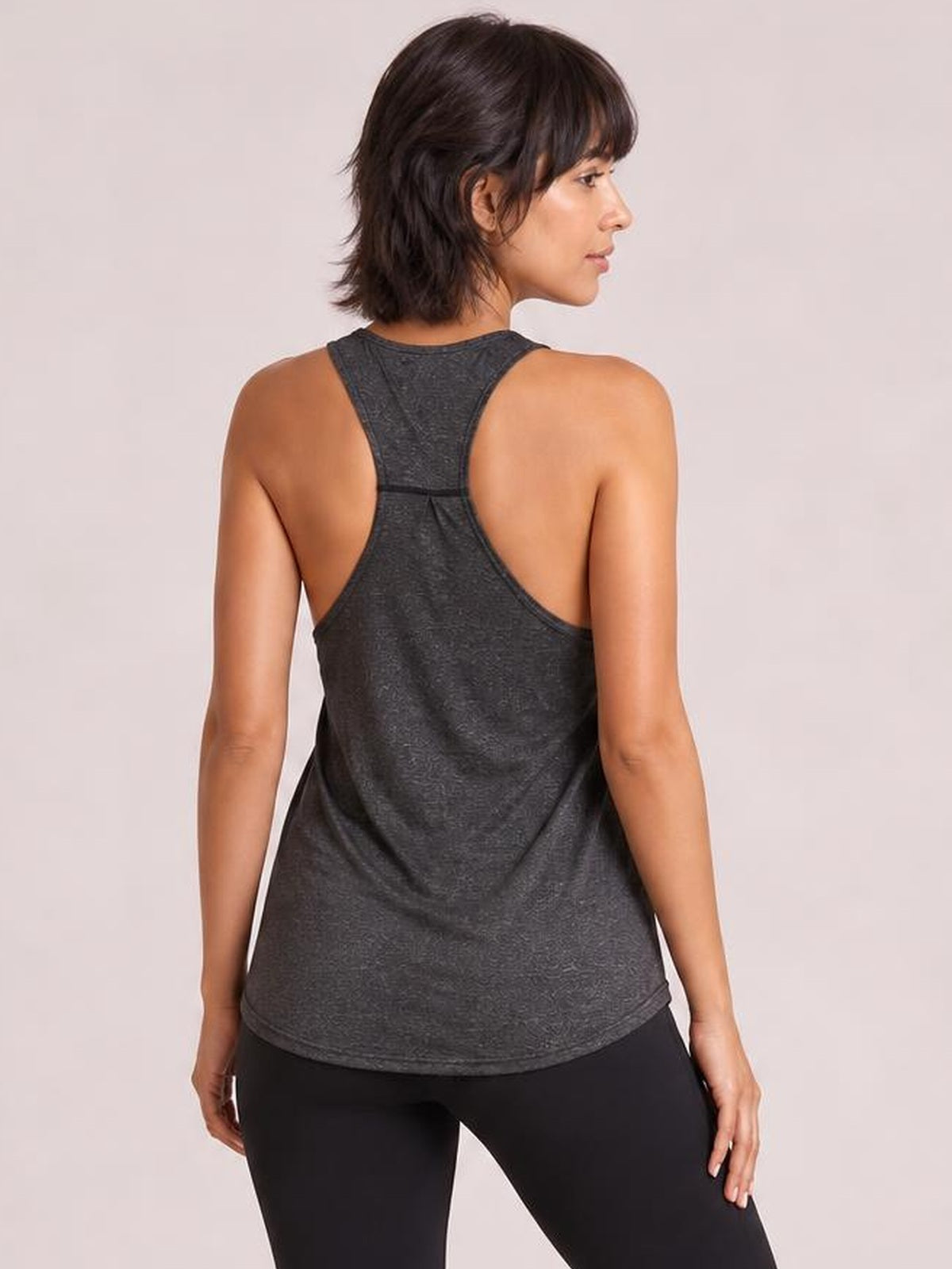 Model poses in a dark gray active tank with wide straps, a racerback construction, open back, and relaxed silhouette over black leggings.