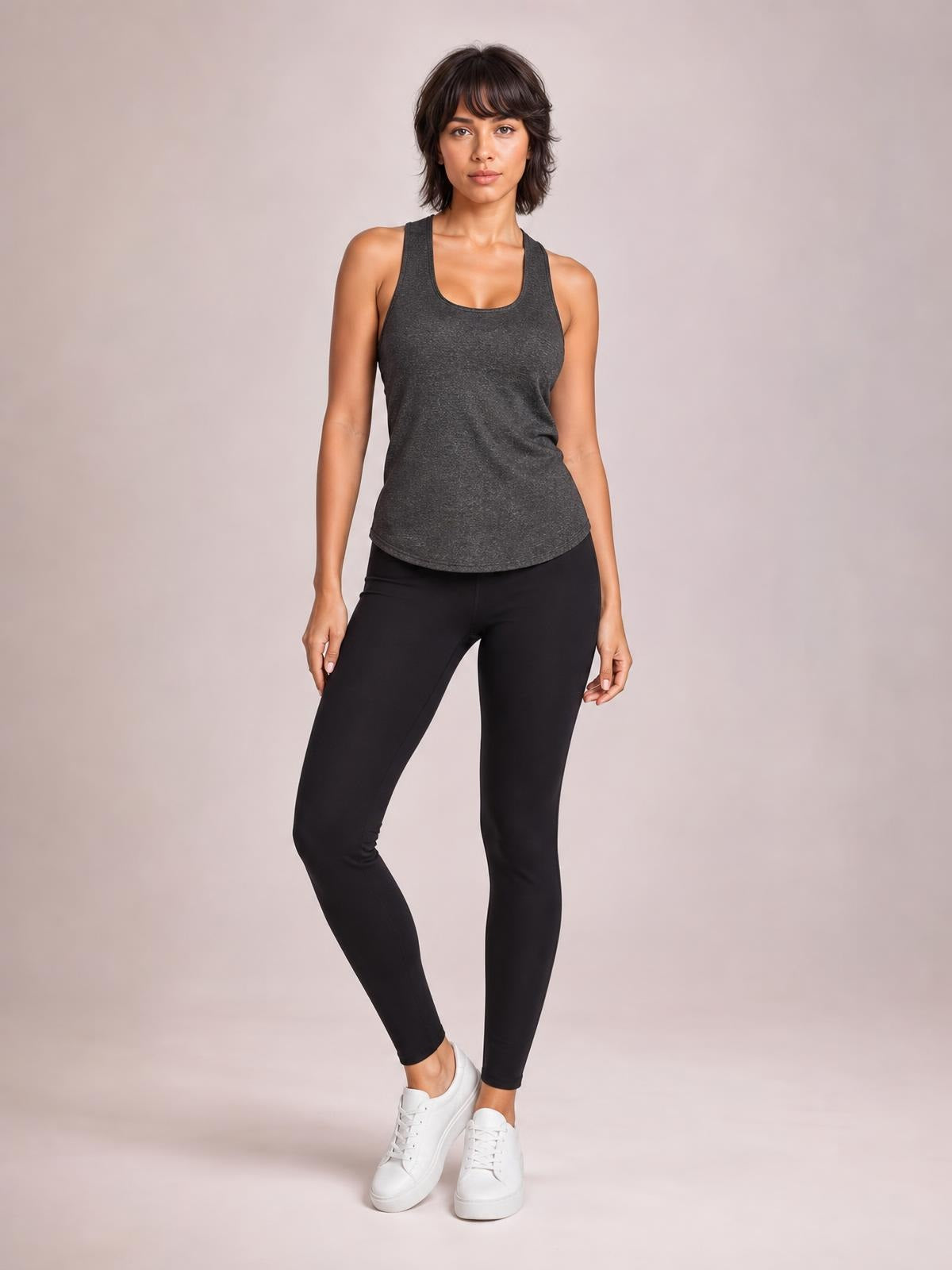 Model poses in a dark gray scoop neck active tank with wide straps, a relaxed straight silhouette, and curved hem, paired with black leggings.