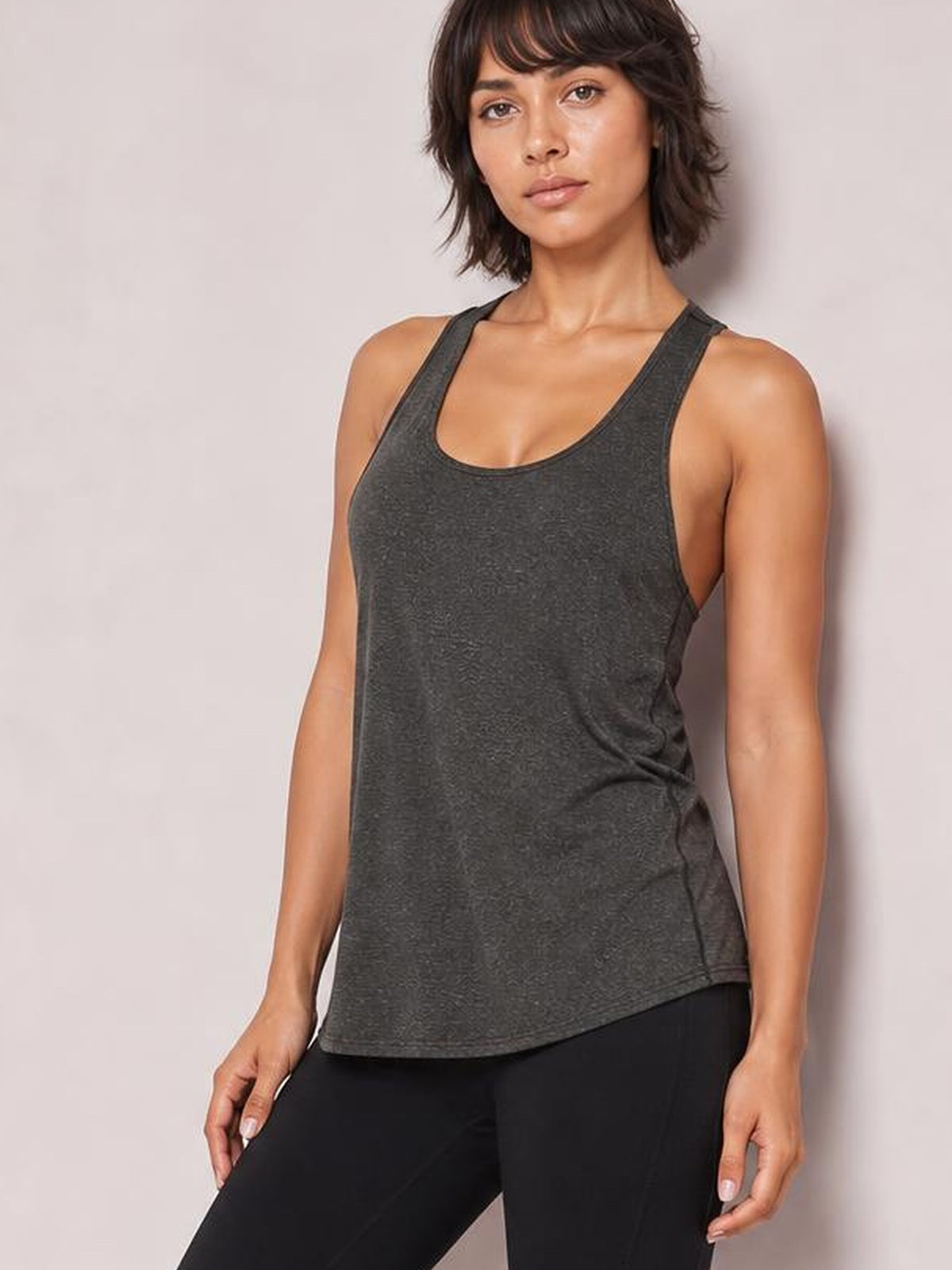 Model wearing a charcoal gray scoop neck active tank with wide straps and a relaxed, slightly curved hem, posed standing with hands at her sides.