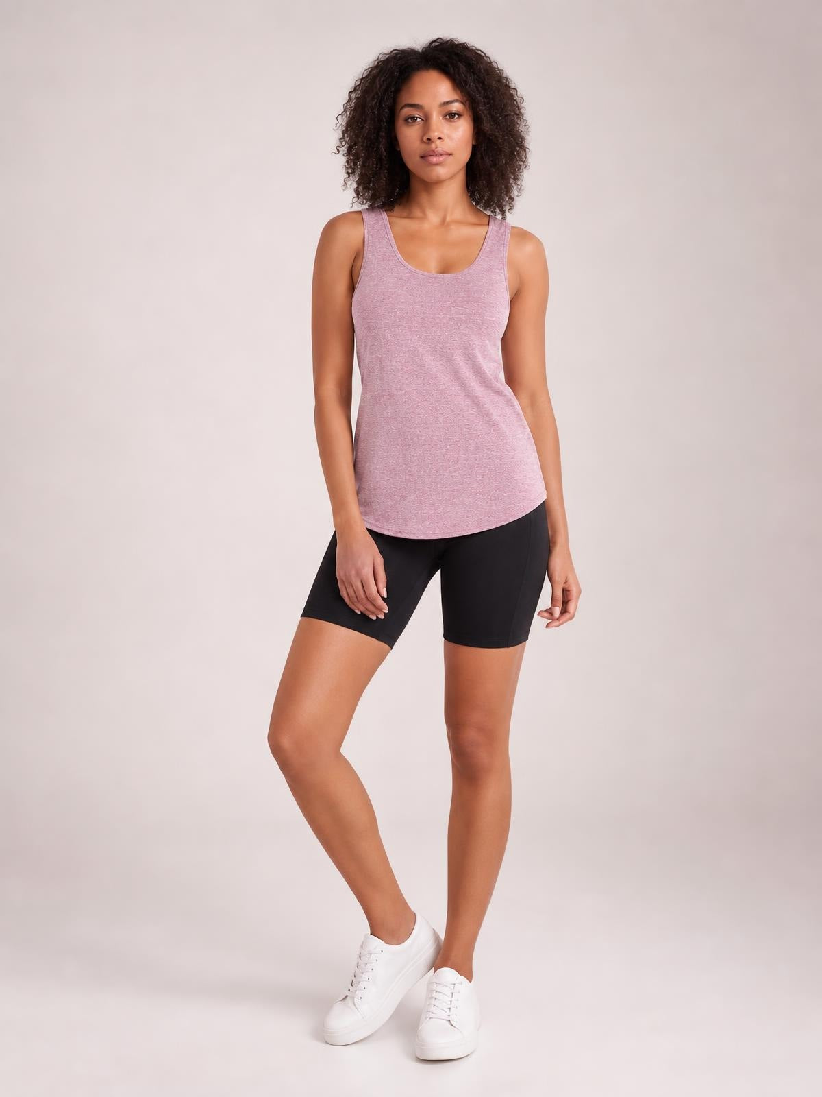Model stands with one leg crossed in a mauve scoop neck active tank featuring wide straps, a fitted silhouette, and a curved hem over black bike shorts.